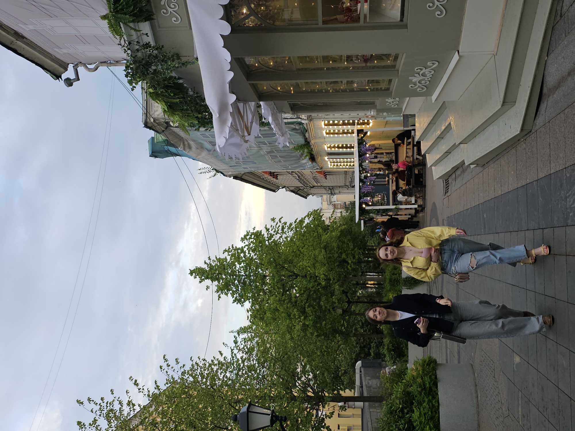Two young women are walking on a cobblestone street in Moscow, Russia, on a cloudy day. The woman on the left is wearing a black blazer over a white t-shirt and gray wide-leg pants, carrying a handbag. The woman on the right is wearing a yellow button-down shirt over a pink top and ripped light-wash jeans, also carrying a handbag. They are walking towards the viewer on a sidewalk lined with trees and shops. One shop has a green facade, white scalloped awnings, and is lit with warm string lights. Another shop, visible further down the street, appears to be a food stall with outdoor seating. The background shows buildings with architectural details and more trees. The overall scene depicts a vibrant urban environment with elements of both nature and commerce.