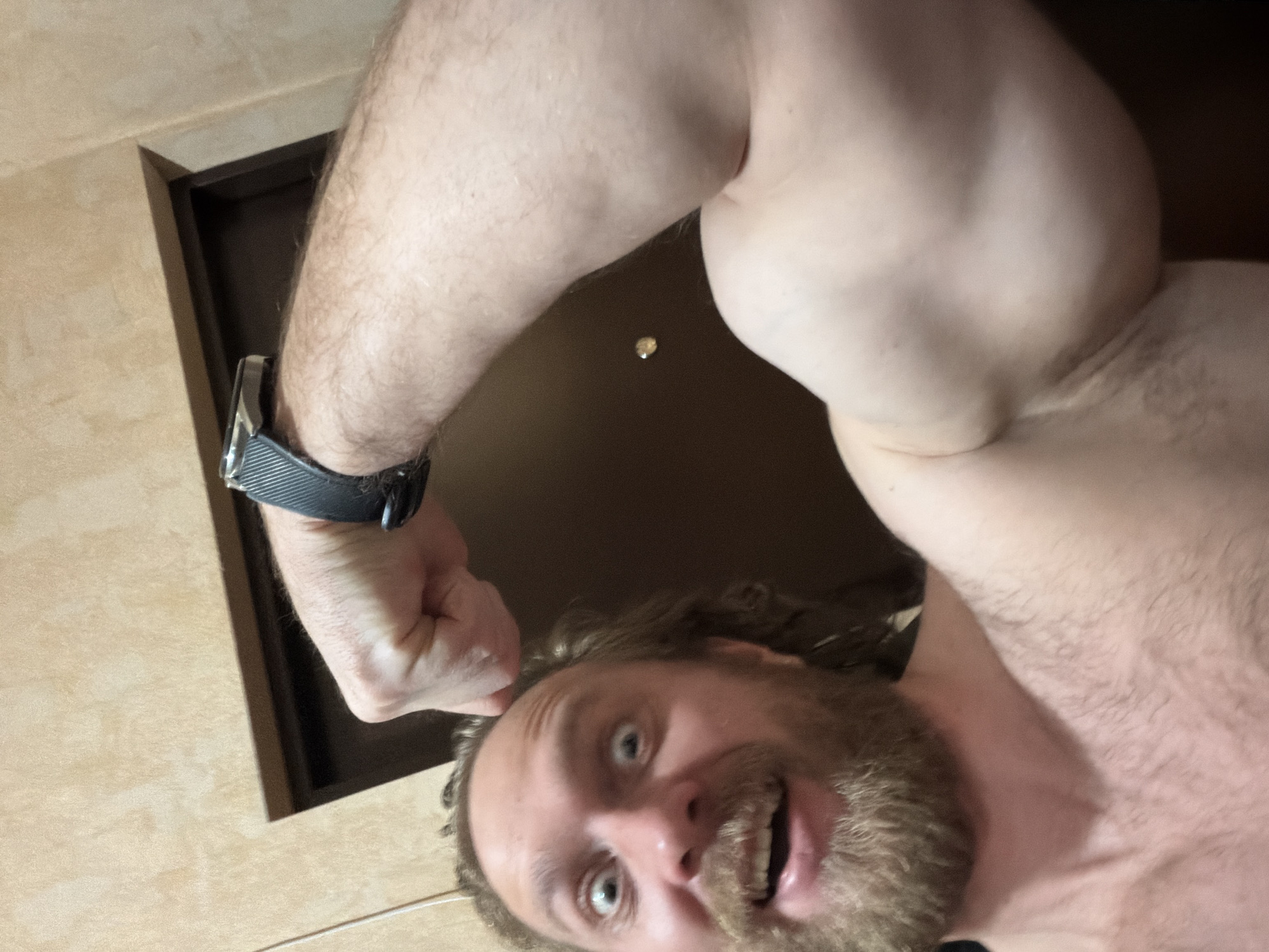 An adult male is featured in a low-angle, close-up selfie, taken indoors, presumably in Saint Petersburg, Russia. The man is shirtless, showcasing his flexed left bicep with a clenched fist. He has a prominent beard and curly, medium-length hair. A dark smartwatch with a textured band is visible on his left wrist.
His facial expression is animated and somewhat exaggerated, with wide, light-colored eyes and an open mouth revealing teeth, suggesting a playful, surprised, or enthusiastic mood. The background consists of a light beige or cream-colored textured wall, possibly tiled, on the left, and a dark brown door frame to the right. A small, circular, shiny object, possibly a light fixture, is visible on the dark surface beyond his arm. The lighting indicates an indoor setting, but the time of day is not discernible. There is no discernible text visible in the image.