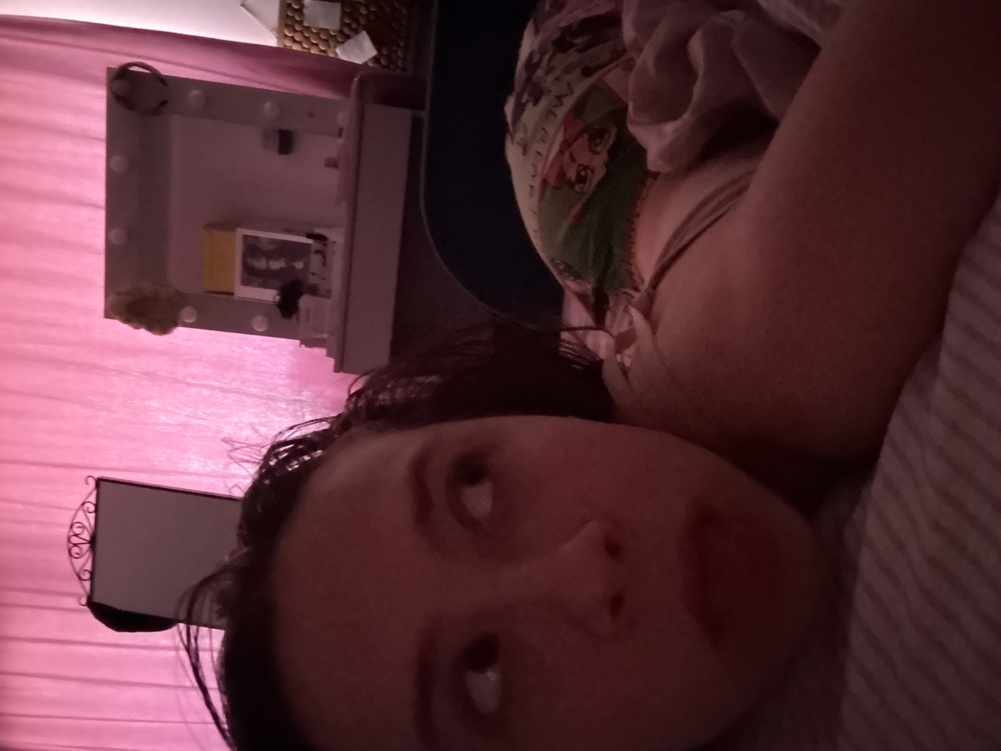 A dimly lit, close-up photograph depicts a person lying down, presumably on a bed, with a patterned mask partially covering their head. The person's face is the primary focus, and they appear to be looking upwards. In the background, elements of a bedroom are visible, including a makeup vanity with a lighted mirror and a wall or curtain with a pink hue. The location of Прикамский, Russia, cannot be determined from the image alone.