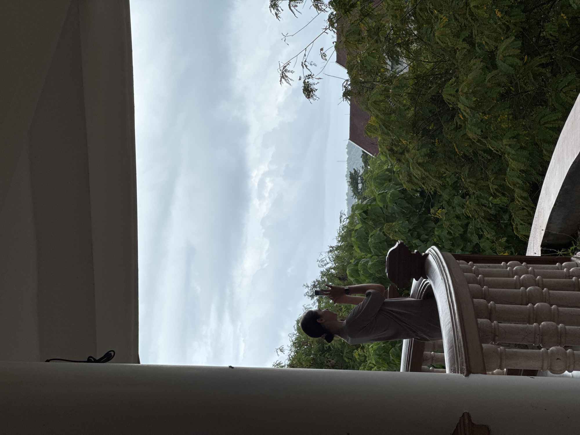A person stands on a balcony in Phuket City Municipality, Thailand, holding a smartphone to take a photo. The person is silhouetted against a cloudy sky and lush green foliage. The balcony has a white railing with decorative balusters. In the background, rooftops of buildings are visible amongst the trees, and a distant hill or mountain range is shrouded in mist. The overall atmosphere is serene and slightly overcast.