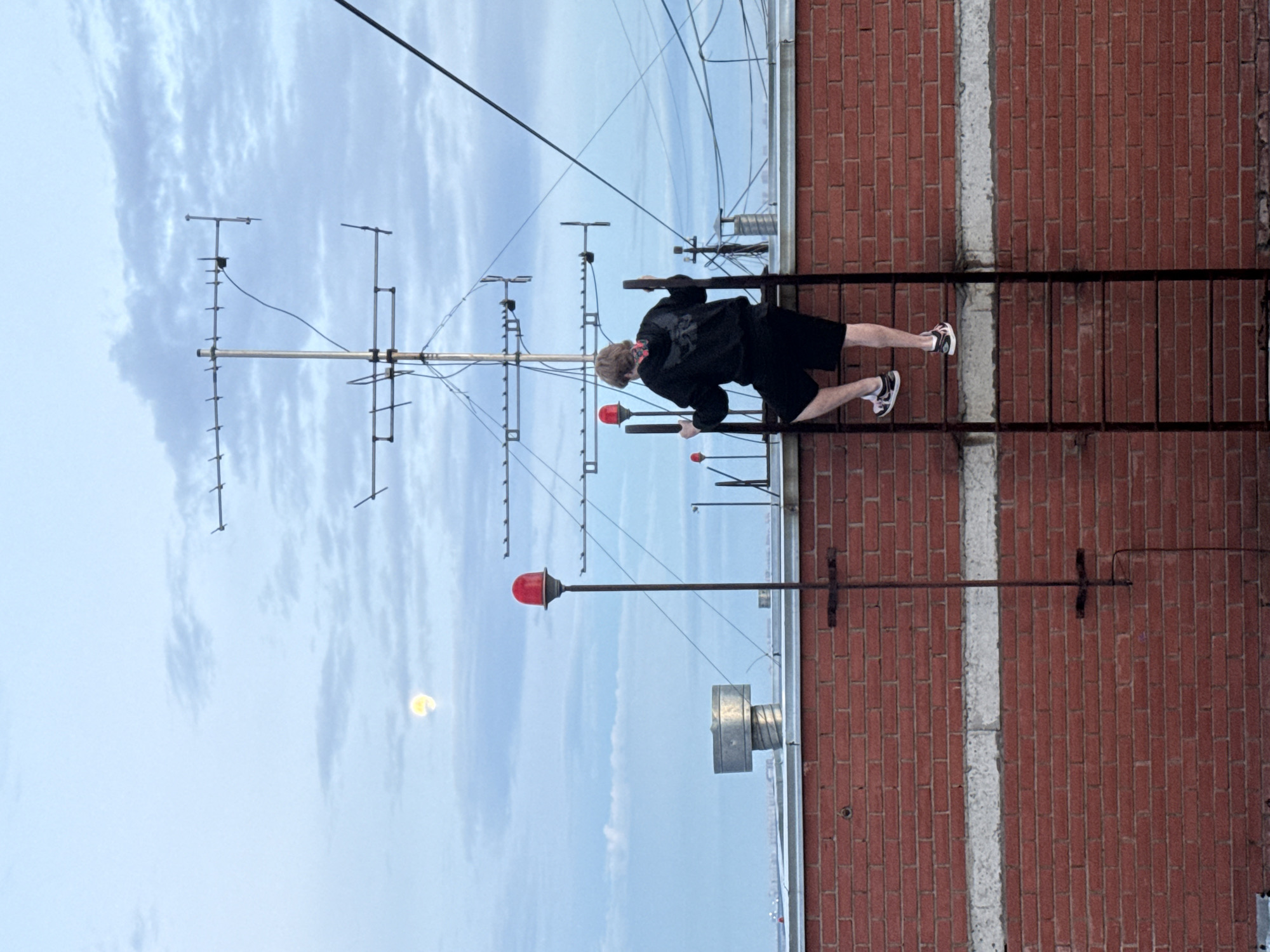 A person is climbing a metal ladder attached to a brick wall. The person is wearing a black hoodie and black shorts. The ladder is on the side of a building in Khimki, Russia. In the background, there are antennas and a cloudy sky. A red light is visible on a pole to the left of the ladder.