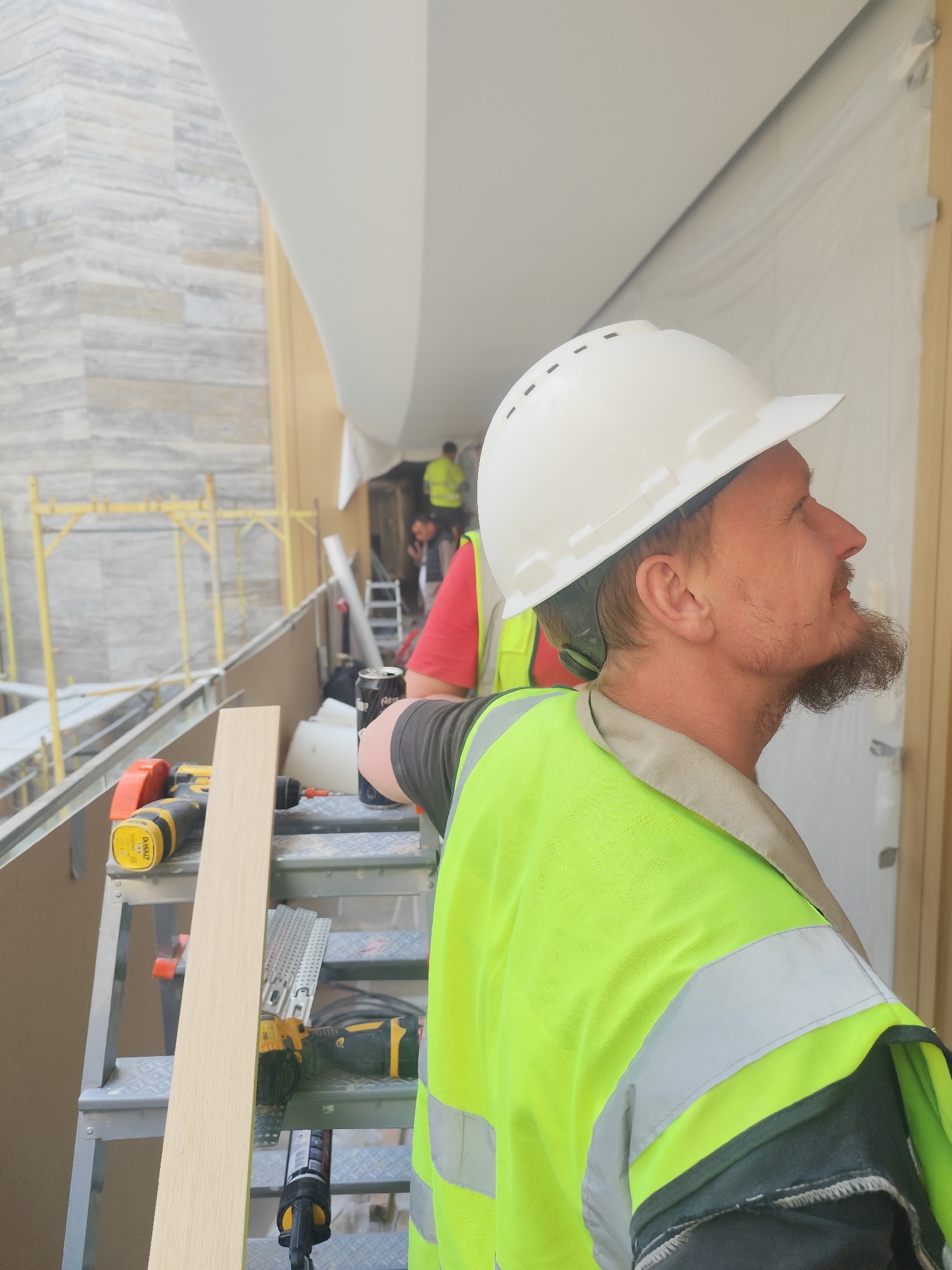This image depicts a construction site during daytime, focusing on a male worker in the foreground, captured in profile. He is looking upwards and to the right, wearing a white hard hat, a neon yellow high-visibility vest with reflective stripes over a dark grey long-sleeved shirt, and a light tan collar. He has a full beard and appears calm and focused.
In the mid-ground, other construction workers are visible, including one in a red shirt and neon yellow vest, and another further back in a bright yellow vest. The background reveals sections of a building under construction, featuring a textured light-colored stone or concrete wall and yellow scaffolding.
On a metal stepladder or platform next to the main worker, several tools and materials are arranged. These include a yellow and black DeWalt DCD771 18V XR Li-Ion drill, a light-colored wooden plank, various metal construction brackets, and a black caulk gun. A black soda can, with "Coca-Cola Zero Sugar" partially visible, is also present.
The immediate environment includes temporary white plastic sheeting or drapery, suggesting ongoing protection or finishing work. A large, curved white architectural element or temporary covering extends above and to the right of the main subject. The scene indicates an active construction or renovation project, likely taking place on an elevated section of a building, such as a balcony or exterior walkway, under diffused natural light.