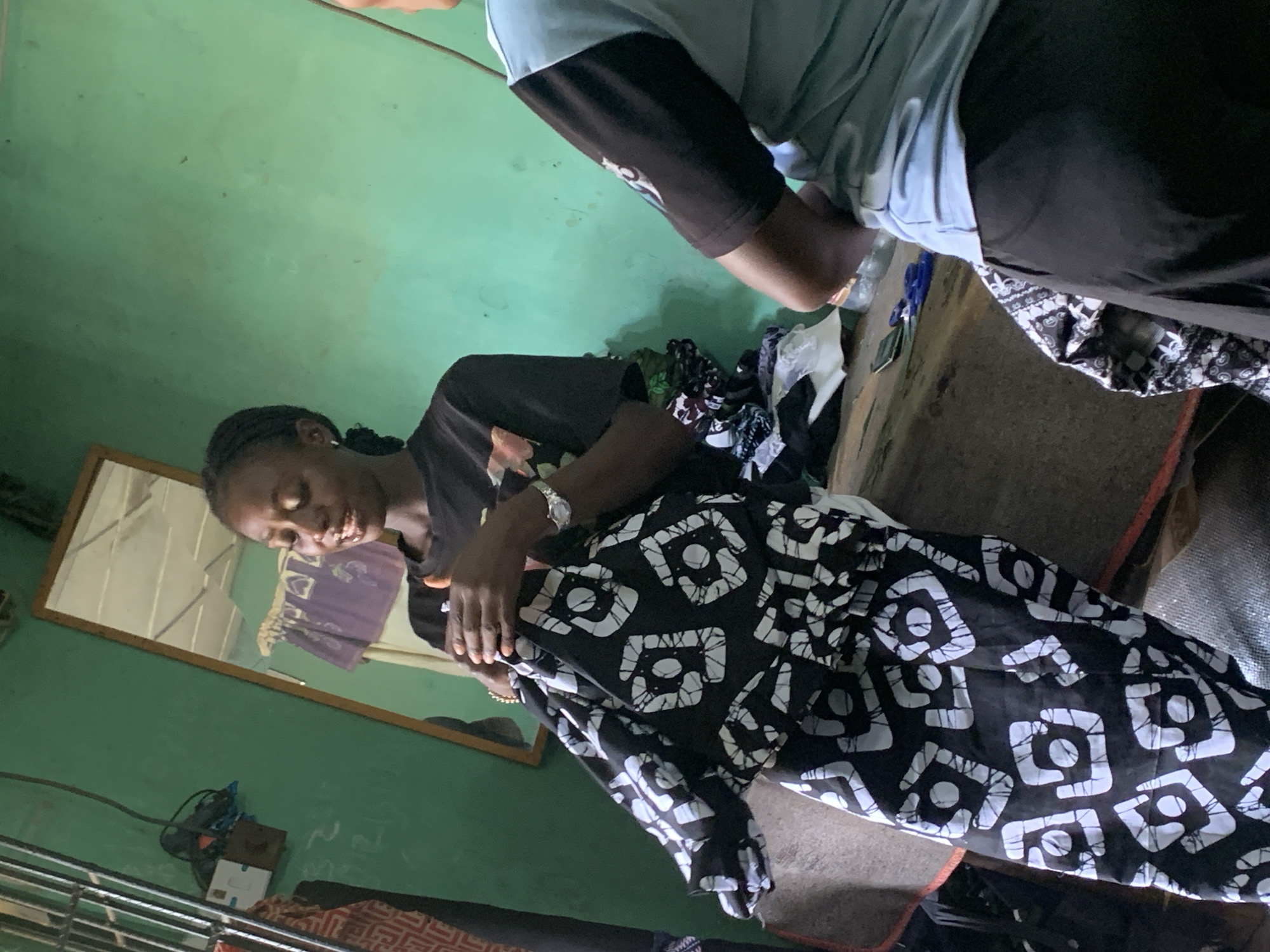 A woman is sitting and holding a black and white patterned garment on her lap. Another person is standing next to her on the right. They are both indoors. The image appears to be taken in Maigizo, Nigeria.