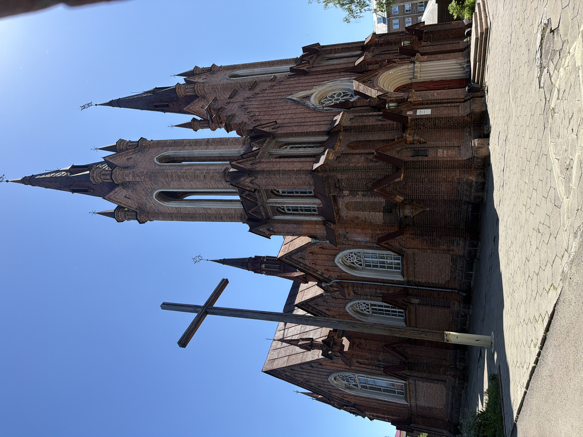 The image shows a red brick church with a tall spired tower, likely in the city of Krasnoyarsk, Russia. A large wooden cross stands in the foreground to the left of the church. The sky is clear and blue. The church has arched windows with decorative tracery and a large rose window above the entrance. The exterior is made of red brick with dark metal accents on the roof and spires. The ground in front of the church is paved with stone tiles. There are no people visible in the image. The scene appears to be a sunny day.