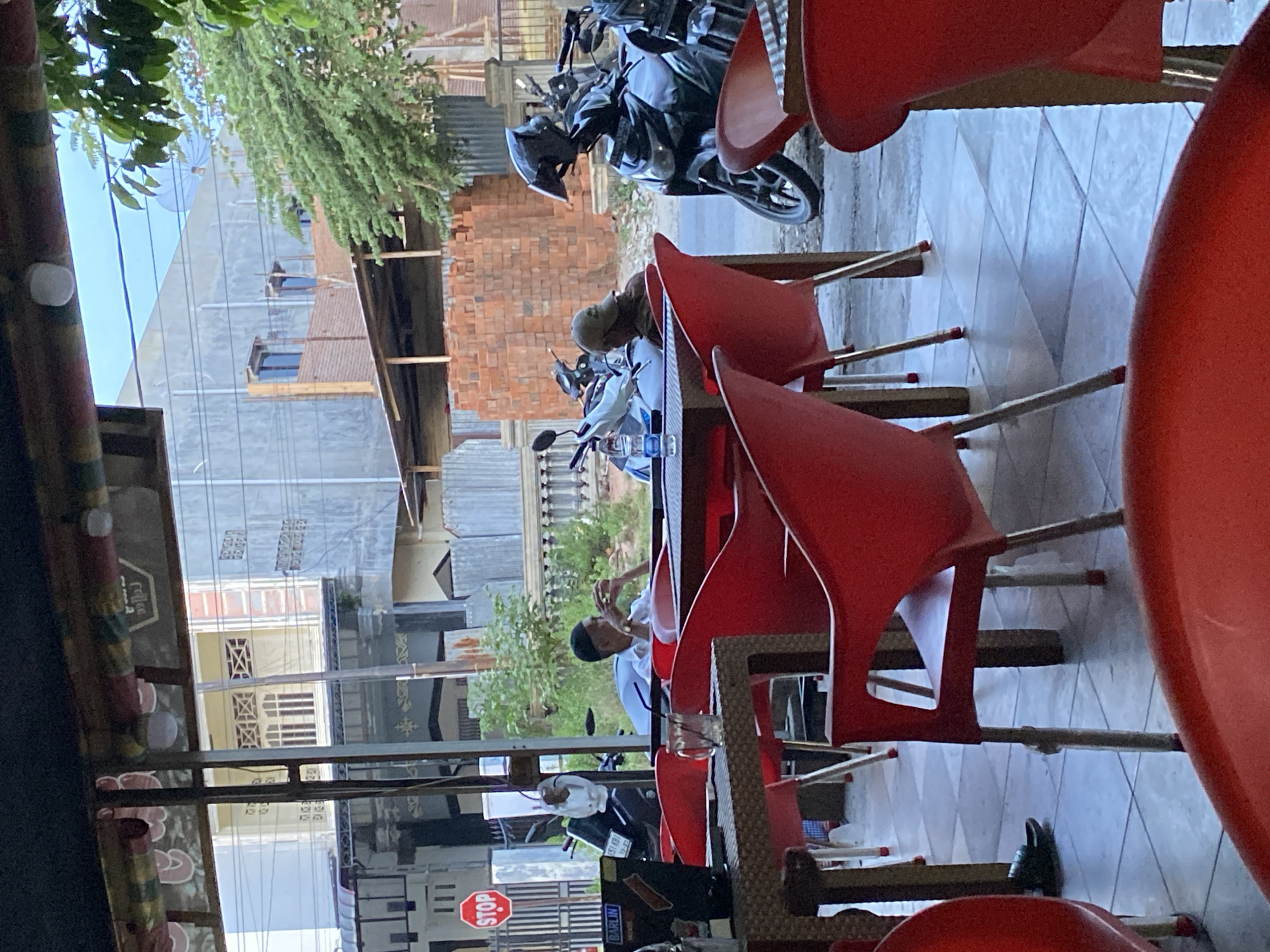 The image captures an outdoor seating area of what appears to be a cafe or restaurant in Lhokseumawe, Indonesia. In the foreground, bright red plastic chairs and tables are arranged on a tiled floor. The chairs have a modern, angular design with metal legs. One table is partially visible in the lower left, and another in the center right.
In the midground, two men are seated at a table. The man on the left is wearing a white shirt and a dark cap, with his hands resting on the table. The man on the right is wearing a brown long-sleeved shirt and a cap, and appears to be looking down at something in his hands. A glass with some liquid is on the table in front of them. Beyond the seating area, several motorcycles are parked.
In the background, the scene opens up to a street view. A red stop sign is visible on the left. Buildings with various architectural styles are seen, including one with a gray facade and a satellite dish, and another under construction with exposed brickwork. Green foliage from trees is interspersed with the buildings. The overall impression is a casual, everyday scene in a Southeast Asian urban setting.