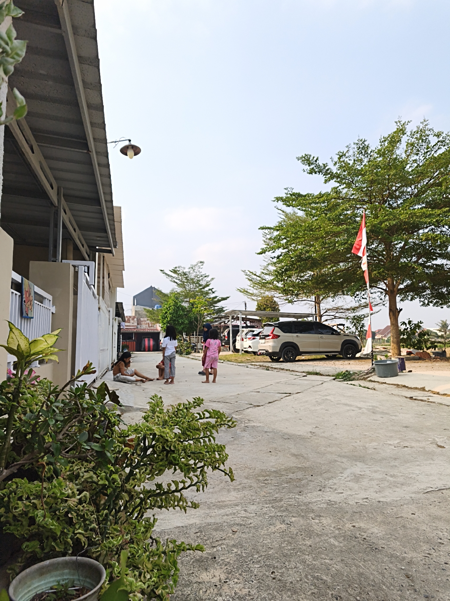 The image depicts a residential street in Bandar Lampung, Indonesia, under a bright, clear sky. In the foreground, a lush green plant in a pot sits to the left, partially obscuring a beige-colored building with a grey, sloped roof. A white gate stands to the right of the building. On the concrete street, several children are present. Two young girls in pink and white clothing are standing, while another child sits on the ground. Further down the street, a light-colored SUV is parked, and a tall tree with vibrant green foliage is visible, adorned with a red and white Indonesian flag. In the background, other residential structures and trees are scattered across the landscape, suggesting a suburban or semi-urban environment. The overall scene conveys a sense of everyday life in a tropical climate.