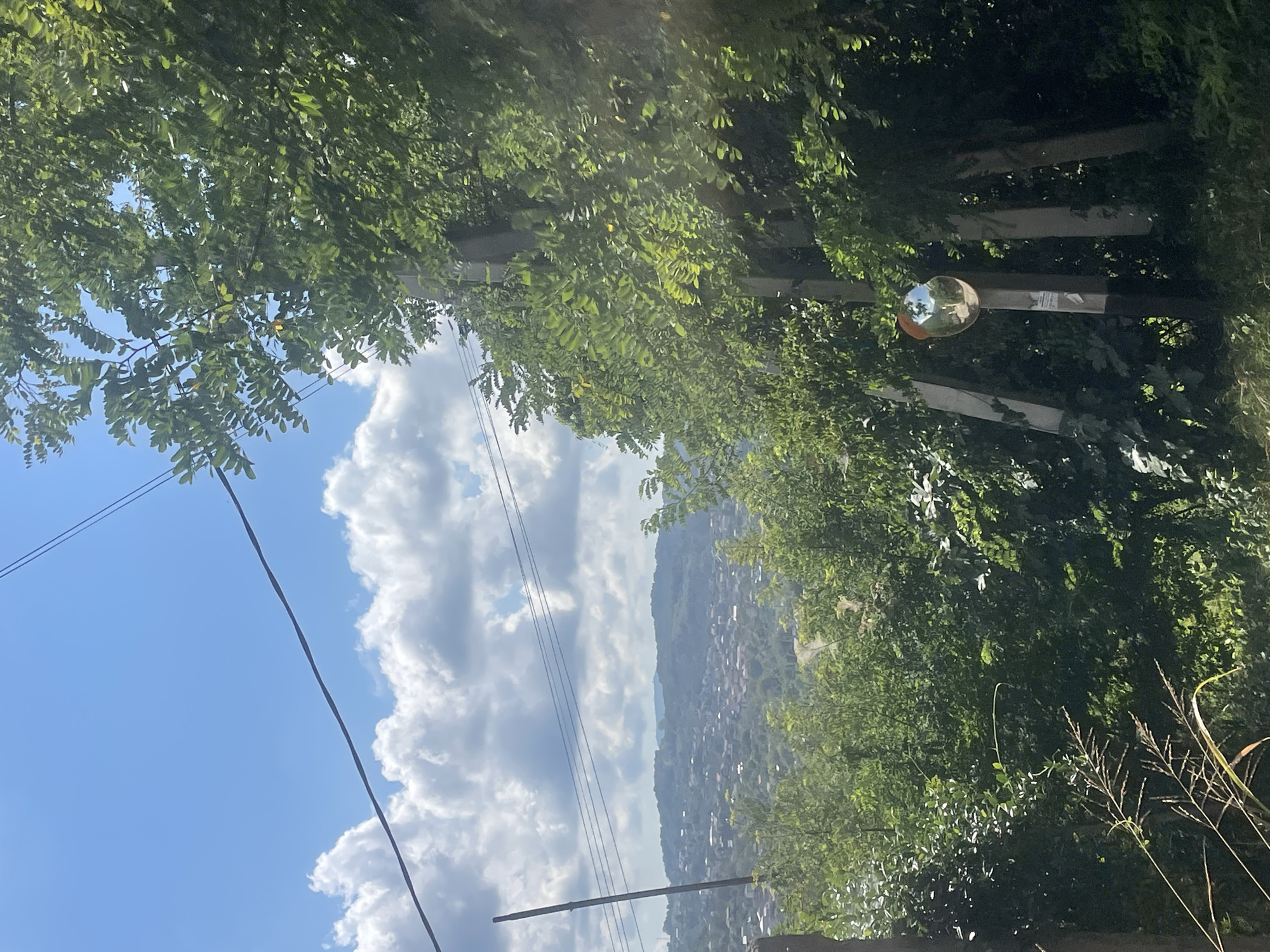 The image captures a scenic view of a hillside town, likely Baranovka, Russia, on a bright, cloudy day. In the foreground, lush green foliage and trees dominate the right side of the frame, with a prominent power pole supporting electrical wires that stretch across the sky. Attached to the pole is a convex mirror, reflecting the sky and a road. Beyond the trees and power pole, the middle ground reveals a densely populated hillside, with numerous houses clustered together and a winding road. The background is characterized by rolling hills covered in verdant vegetation, extending into the distance under a sky filled with puffy white clouds. The overall impression is one of a rural or semi-rural setting bathed in natural light.