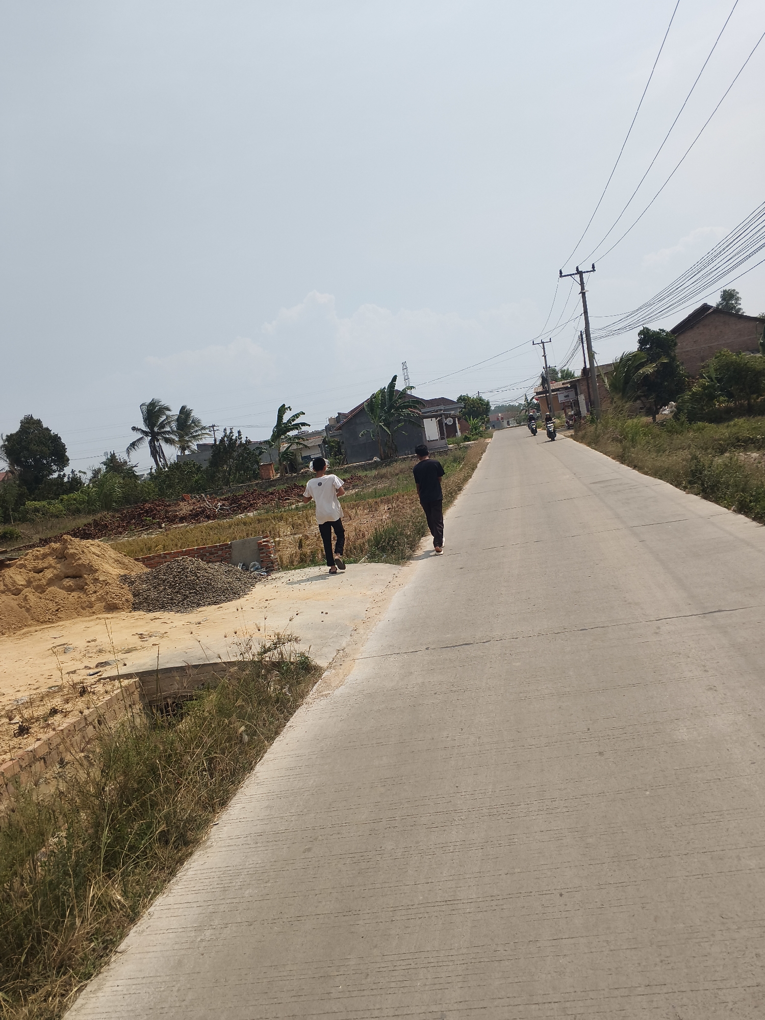In Banjar Agung, Indonesia, a scene unfolds on a paved road bordered by undeveloped land. Two individuals are present. One, a male wearing a white t-shirt and dark pants, walks away from the viewer along the right edge of the road. Another male, dressed in a black t-shirt and dark pants, walks further down the road on the left side. The road itself is cracked and shows signs of wear. The undeveloped land to the left features piles of sand and gravel, brick construction materials, and dry grass. To the right of the road, houses with dark tiled roofs and various types of vegetation, including palm trees and leafy trees, are visible. Utility poles and overhead power lines stretch across the sky, which is bright and hazy. Two motorcycles are seen in the distance on the road. The overall environment suggests a semi-rural or developing urban area under a clear, sunlit sky.