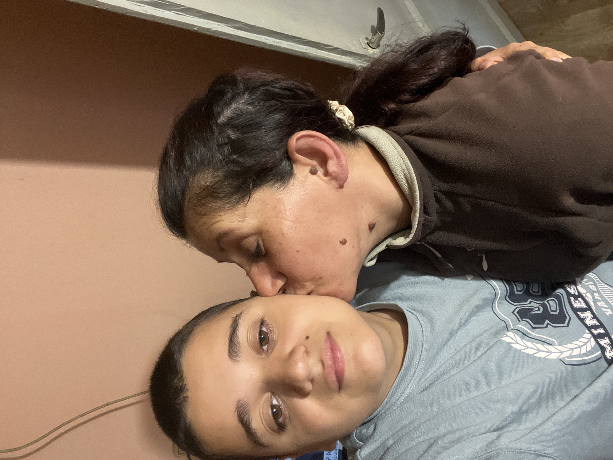 The image shows two individuals. One person with dark hair is kissing the forehead of another person wearing a blue t-shirt. The room has peach-colored walls. The location is Lozarevo, Bulgaria.