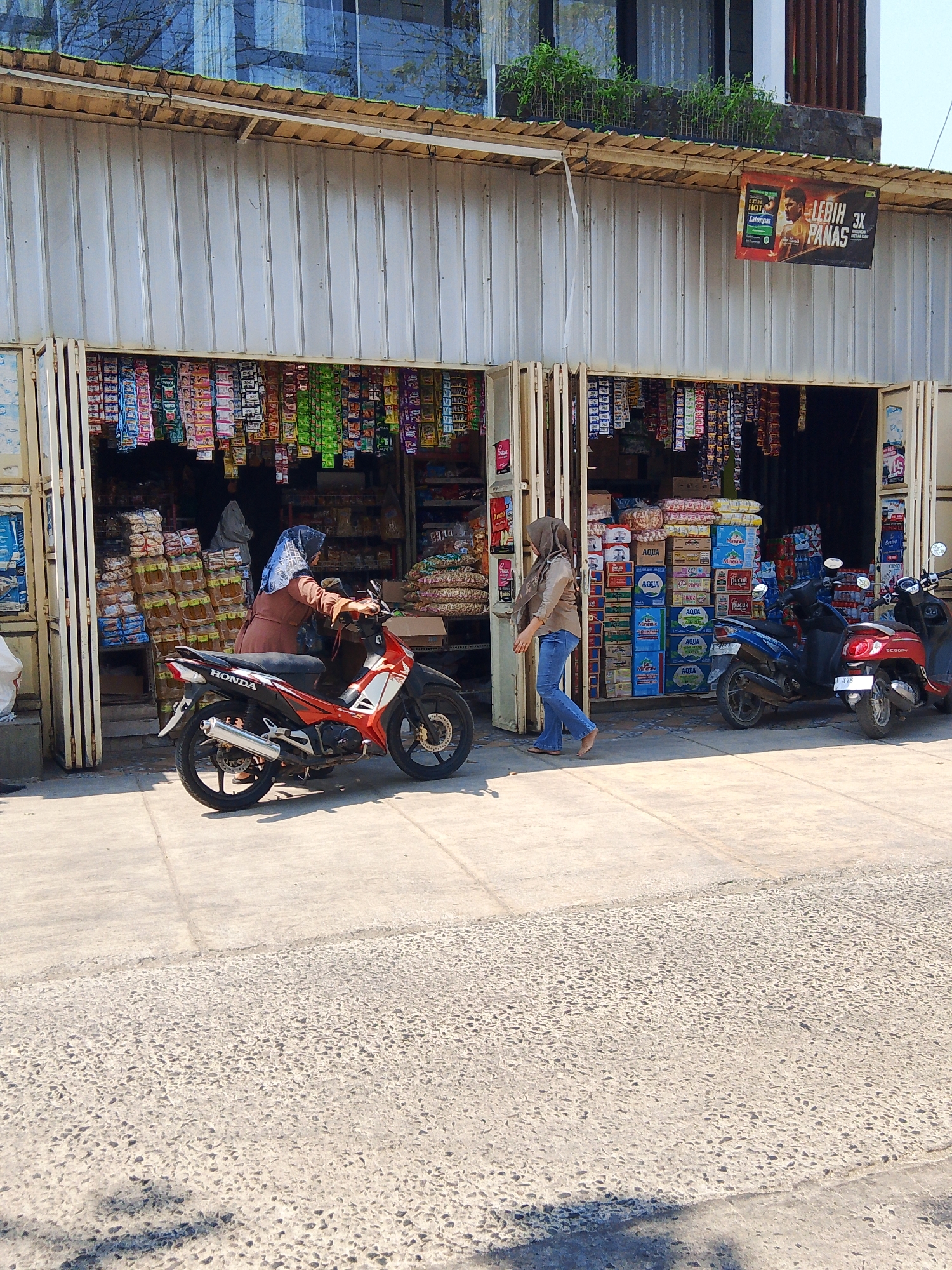 A storefront in Kedondong, Indonesia, features a corrugated metal facade with various goods displayed. The store appears to be a small convenience store or warung, with shelves stocked with packaged items such as snacks, drinks, and household goods. Two women are present; one is near a motorcycle parked in front of the store, and the other is walking away from the store. Several motorcycles are parked along the sidewalk in front of the store, suggesting a busy commercial area. The building behind the storefront has windows reflecting the sky and surrounding trees. The foreground shows a paved sidewalk and a gravel road.