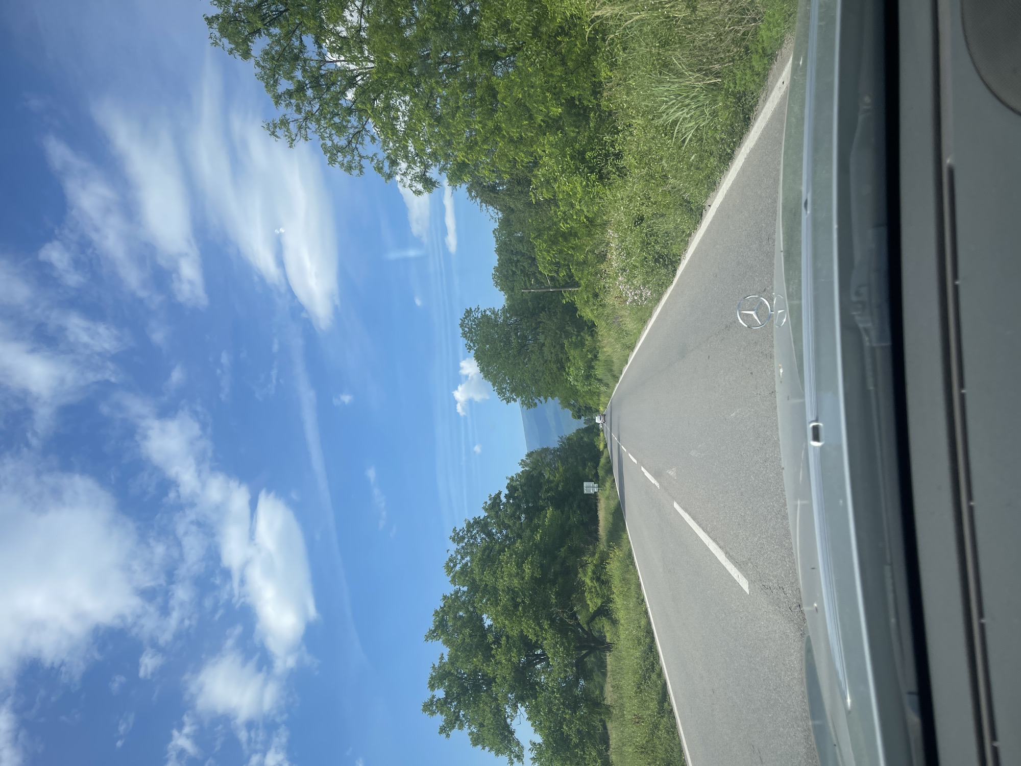The image shows a perspective from inside a car driving on a rural road. The road is paved, with a white line dividing lanes. On either side of the road are grassy verges and lush green trees. The sky is bright blue with scattered white clouds. In the distance, a mountain range is visible. The hood of the car in the foreground features a Mercedes-Benz emblem. There are no people visible in the image. The scene is consistent with a drive through the countryside near Mezdreya, Bulgaria, given the presence of rural roads, vegetation, and distant mountains typical of the region.
