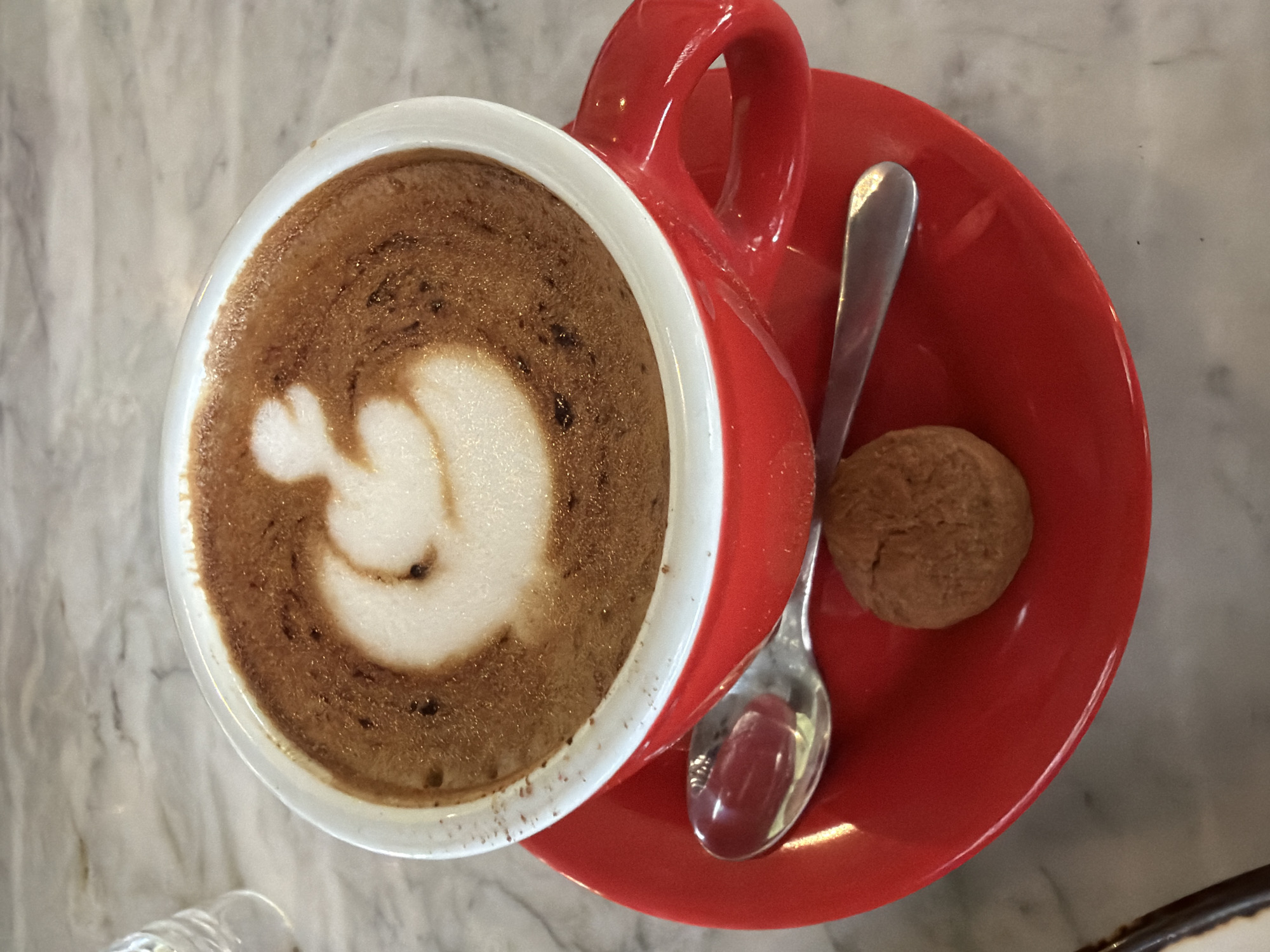 A high-angle shot reveals a red ceramic coffee cup and saucer set placed on a light gray marble surface. The coffee cup is filled with cappuccino or latte art in the shape of a figure. A small, round cookie and a silver spoon are positioned on the saucer next to the cup. The location is North Kuta, Indonesia.