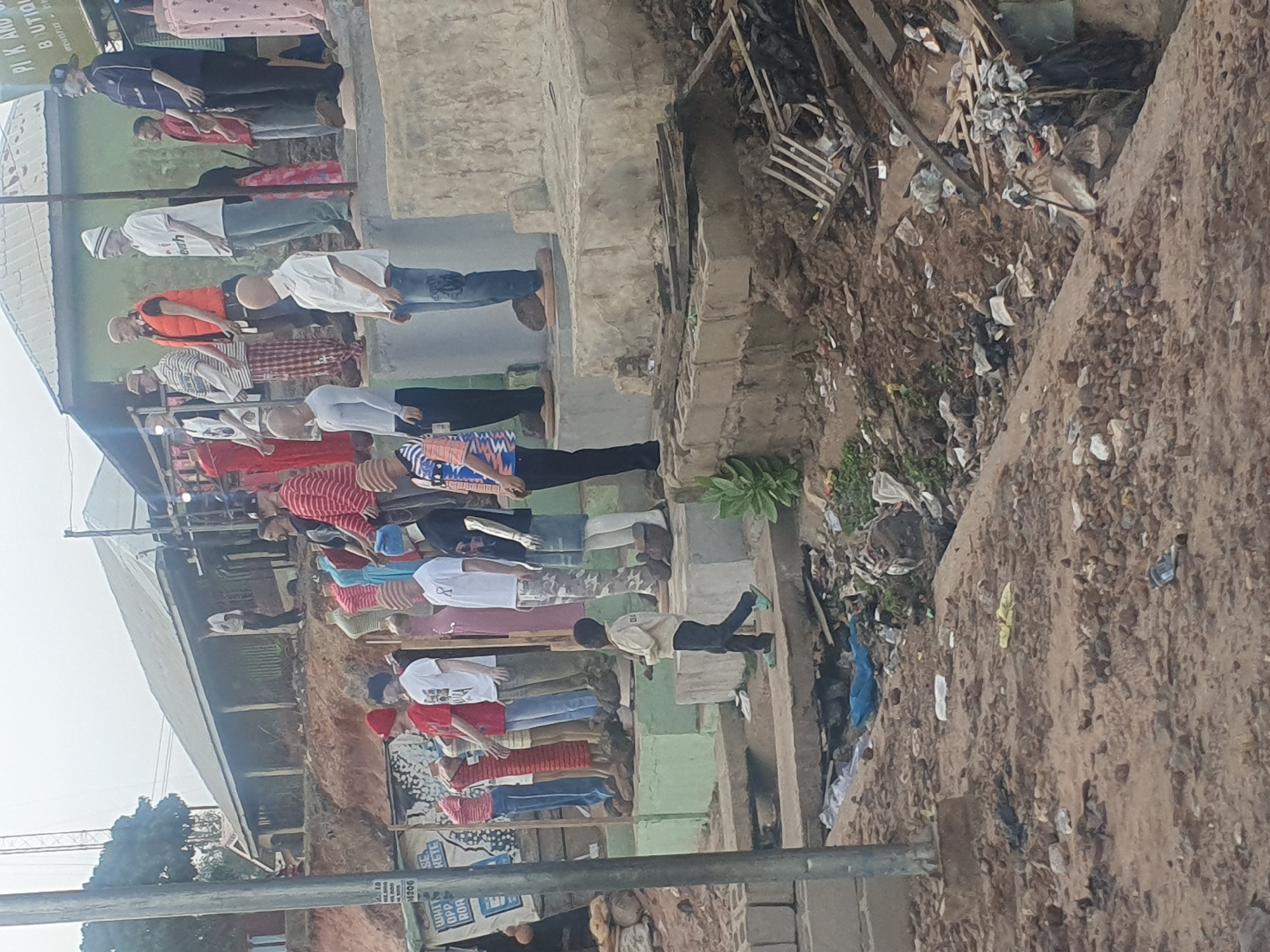 In Lokoja, Nigeria, a group of approximately 15 people stand in a line along a raised, concrete walkway. The line extends to the left side of the frame. Individuals are diverse in dress, wearing items such as t-shirts, patterned shirts, and dresses, with some wearing hats or masks. An open drainage ditch runs parallel to the walkway, filled with refuse. Beyond the ditch, there is a dirt area with scattered debris. The walkway is adjacent to a building with a grey roof. A utility pole is visible in the lower left corner.