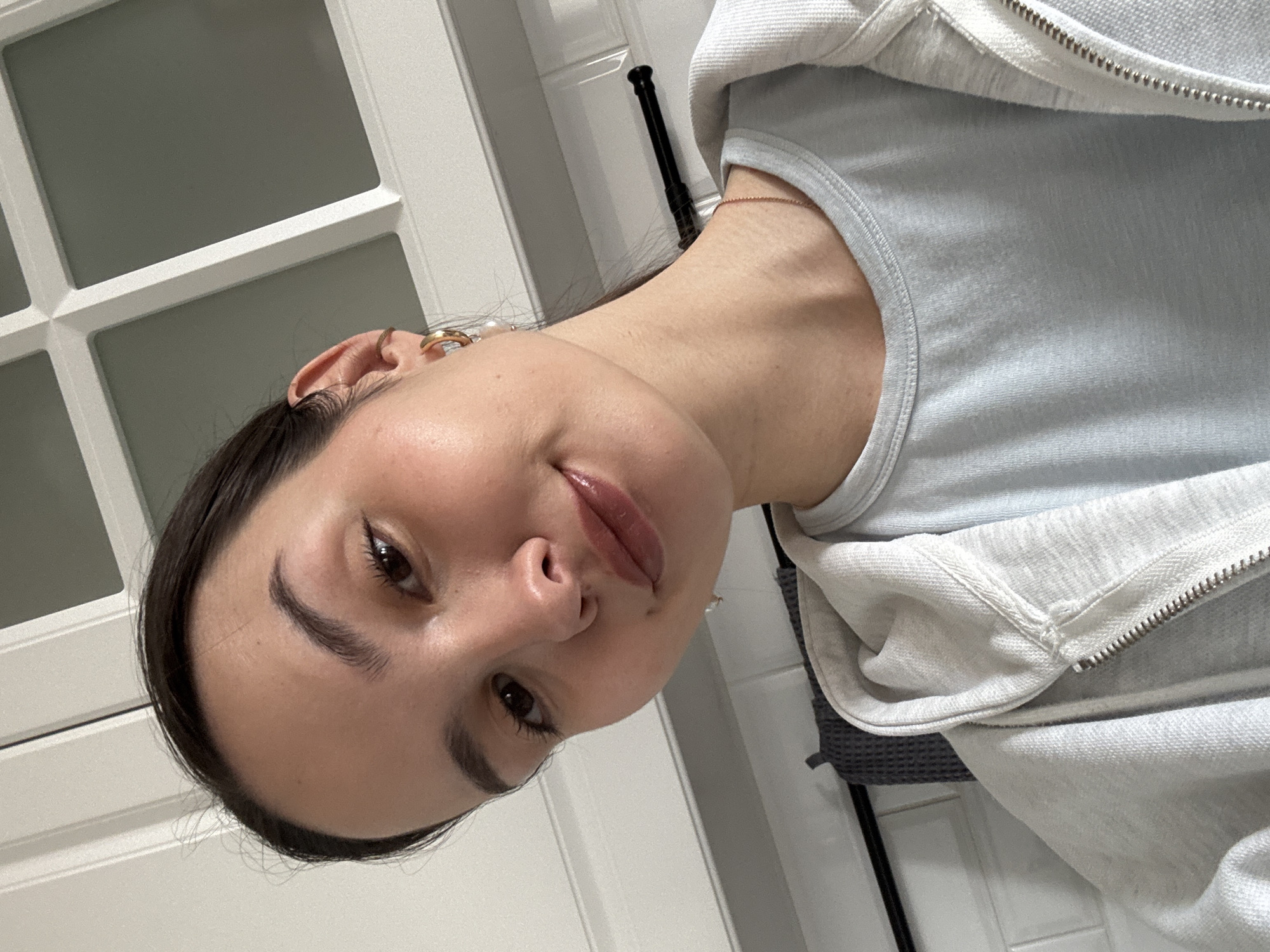 A casual portrait features a young woman, likely in Chaikovsky, Russia, taking a selfie indoors. She is positioned with her head tilted, looking directly at the camera with a subtle, pleasant smile and parted, glossy lips. Her dark hair is pulled back, highlighting her dark eyes and well-groomed eyebrows.
She is wearing a light blue or pale grey tank top, layered under a light grey hooded sweatshirt with a silver zipper. A delicate rose gold chain necklace is visible around her neck, and she sports gold hoop earrings and a smaller gold stud in her left ear.
The background consists of light-colored, possibly white, paneled doors or wall sections, suggesting an interior setting like a bedroom or bathroom. A dark, vertical object, possibly a handle or hook, is visible on the right side of the frame. The lighting is soft and even, indicating an indoor environment, likely illuminated by artificial light. There is no visible text in the image.