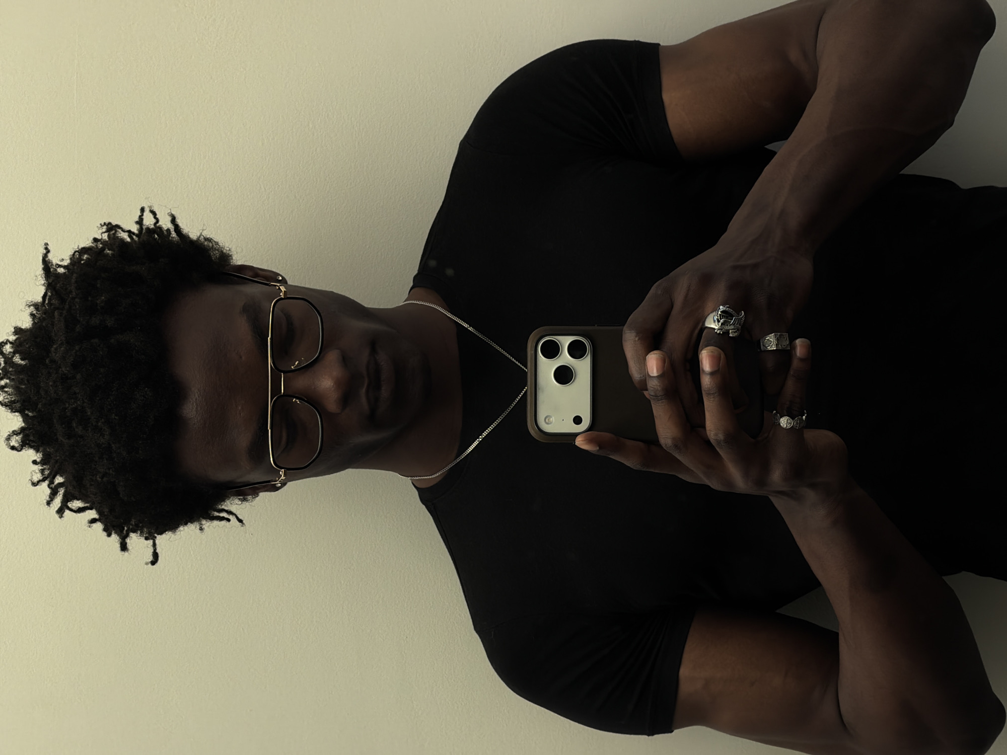 A man is taking a selfie indoors against a plain, light-colored wall. He is wearing glasses, a black t-shirt, and a silver necklace. He has short, curly hair. The phone in his hands is dark-colored with three camera lenses. The man has rings on his fingers. The location is not identifiable as being in London, United Kingdom.