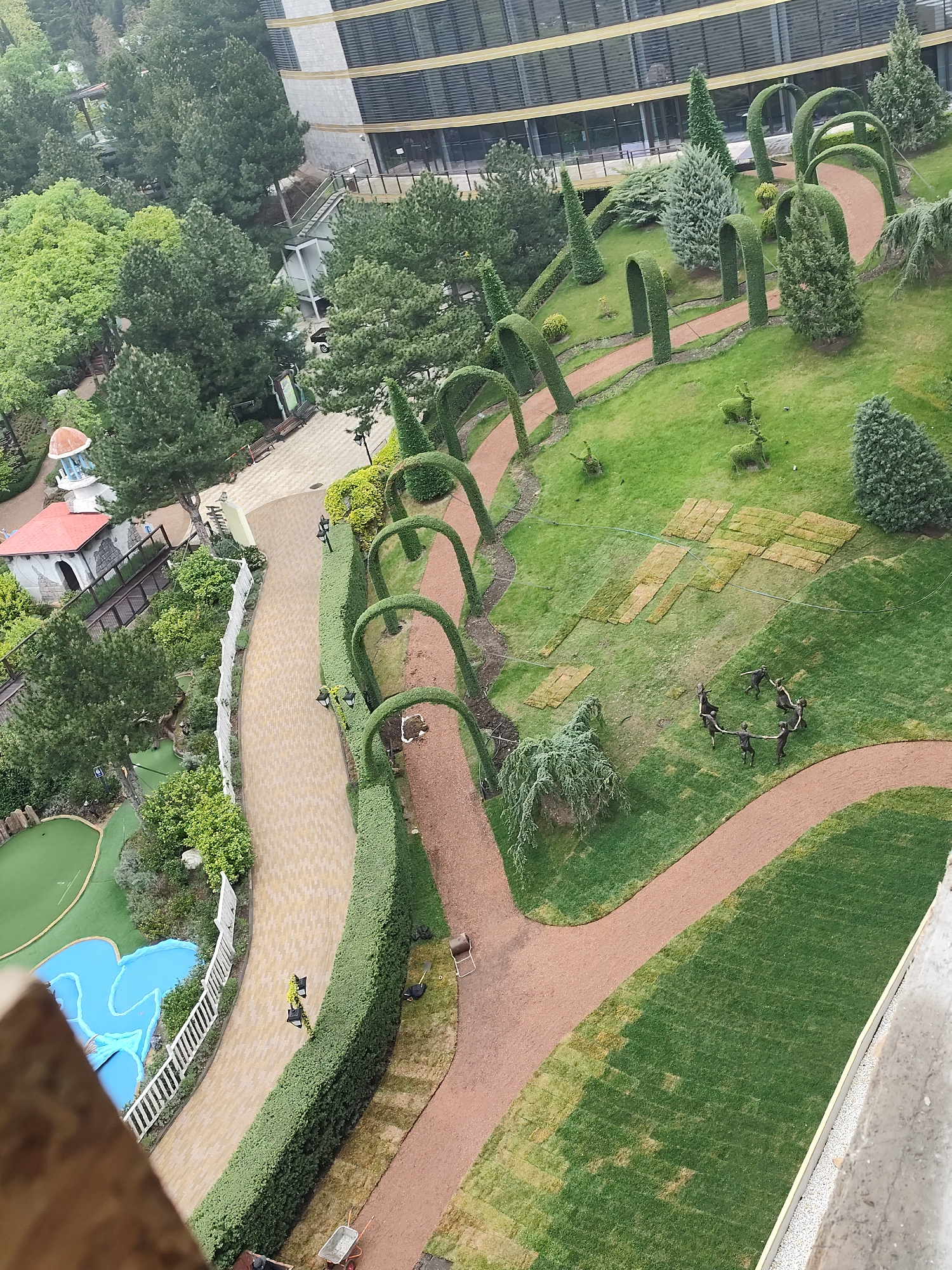 An aerial view shows a landscaped park in Симеизский поселковый совет, Ukraine. The park features winding pathways of reddish-brown and tan hues. Sculpted hedges and trees line the paths, including arched hedge structures. There's a mini-golf area with blue water features enclosed by a white fence. Additional details include topiary animal figures on the lawn, grass sections laid out like puzzle pieces, and a circle of human-shaped figures. A building with a multi-story facade is visible in the background.
