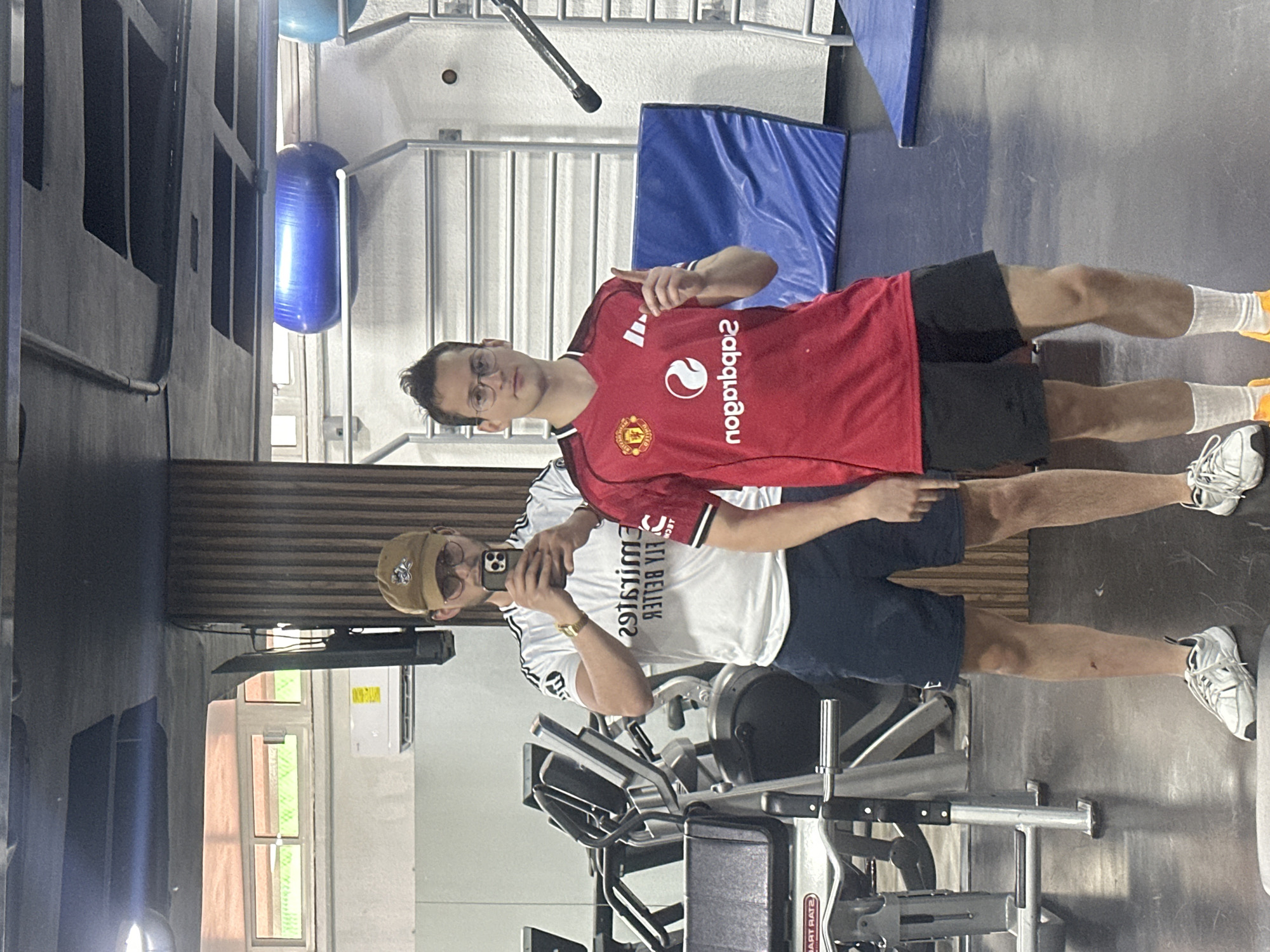 Two men are standing in a gym, taking a selfie in a mirror. One man is wearing a red Manchester United soccer jersey and black shorts. The other man is wearing a white soccer jersey, navy shorts, and a tan baseball cap. A weight machine is in the foreground. Gym equipment is scattered around. The location is the city of Huixquilucan de Degollado, Mexico.