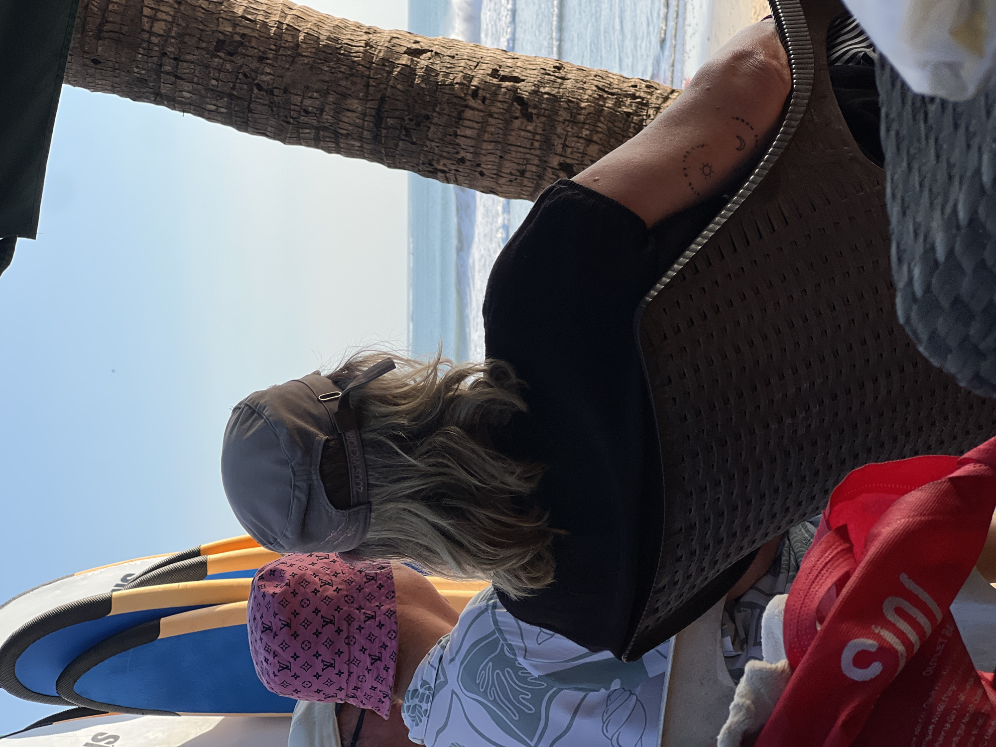 The image shows a rear view of two individuals sitting on chairs facing a beach and ocean. To the left, a person wears a pink bucket hat with a monogram pattern and a patterned shirt. The person to the right has long blonde hair, wears a gray baseball cap, and a black t-shirt. Their right arm is visible, showing a tattoo of a sun and moon design. Behind them, several surfboards are propped up. A palm tree trunk is visible on the right side of the frame, and a dark green umbrella overhangs the top. The ocean has moderate waves, and the sky is clear and bright. The overall scene depicts a relaxed setting at a beachside location. The presence of surfboards and the tropical vegetation suggest a location like Kuta, Indonesia, a popular tourist destination known for its beaches and surf culture.