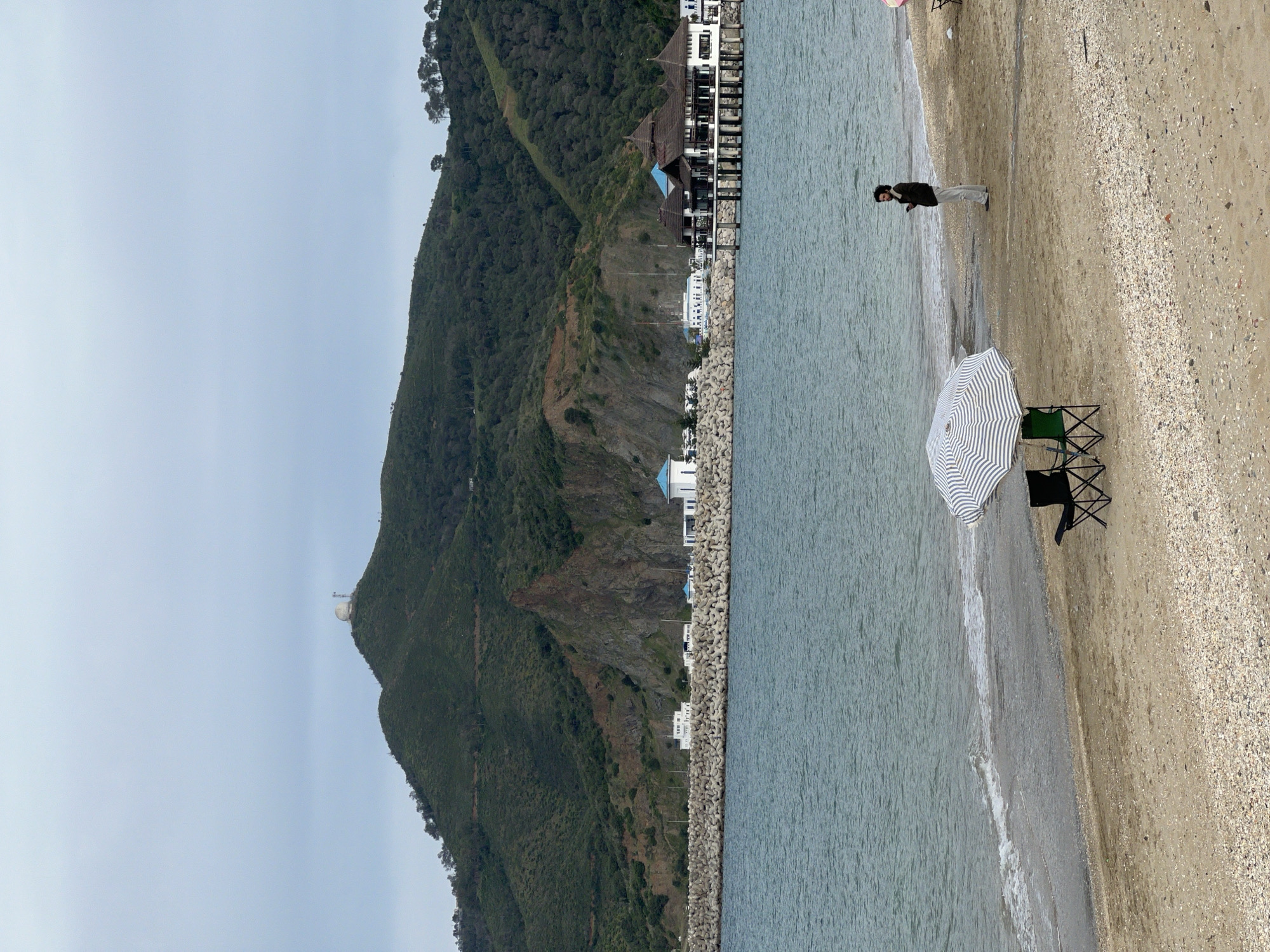 The image captures a beach scene in M'Diq, Morocco. A mountain covered in green vegetation dominates the left side, with a white structure visible at its peak. Along the base of the mountain, there are buildings and a protective barrier made of large concrete blocks separating the mountain from the beach and the sea.
On the right side of the image is a sandy beach, with a striped umbrella and two dark-colored folding chairs set up near the water's edge. A person is walking along the shoreline, wearing dark-colored clothing and light pants. The sea is calm, with small waves gently washing onto the shore. The sky is overcast and gray.