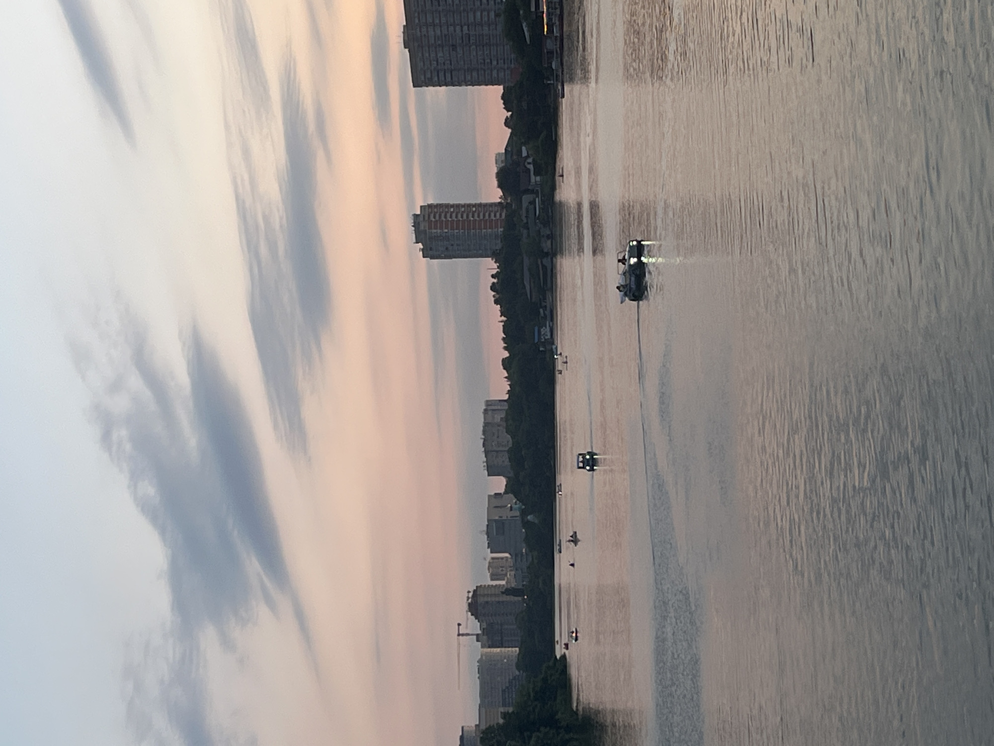The image depicts a serene sunset over the Kuban River in Krasnodar, Russia. The sky is painted with soft hues of pink and lavender, with wispy clouds stretching across the horizon. In the foreground, the river reflects the tranquil sky, with ripples disturbing the otherwise glassy surface. Several small boats are visible on the water, some appearing to be paddle boats, while others are motorboats. One motorboat in the middle right of the frame is illuminated by its headlights, suggesting it is moving. In the background, the cityscape of Krasnodar is visible, characterized by a mix of residential buildings and what appear to be construction cranes, indicating ongoing development. Trees line the riverbank, providing a lush green contrast to the urban structures. The overall atmosphere is one of peaceful coexistence between nature and urban life at dusk.