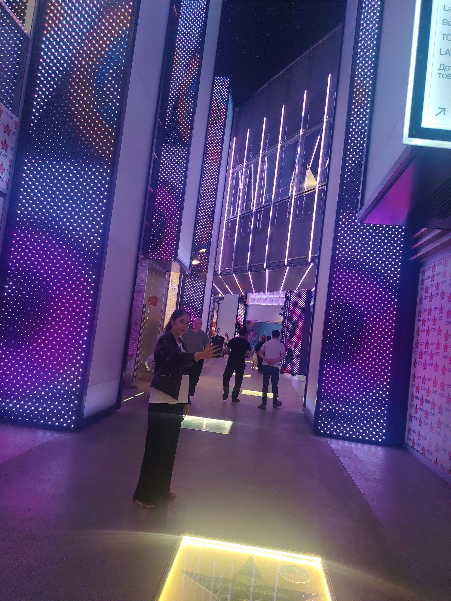 The image depicts an indoor promenade illuminated by extensive LED light displays. The walls are adorned with panels of small, multicolored LEDs, predominantly in shades of purple and blue, creating a vibrant, futuristic atmosphere. Vertical rows of thin, bright pink LED lights run along a section of the architecture. Several individuals are present, walking through the space. In the foreground, a woman is taking a selfie with her phone. Other people are seen in the background, some walking in groups, others individually. The floor is a smooth, dark surface with embedded light panels that emit a yellowish glow. The overall environment suggests a modern entertainment or commercial venue, possibly a shopping mall or an entertainment complex, characterized by its dynamic light installations. There is no visible indication of the specific location being within the city of Sovkhoza imeni Lenina, Russia, or any other city for that matter.