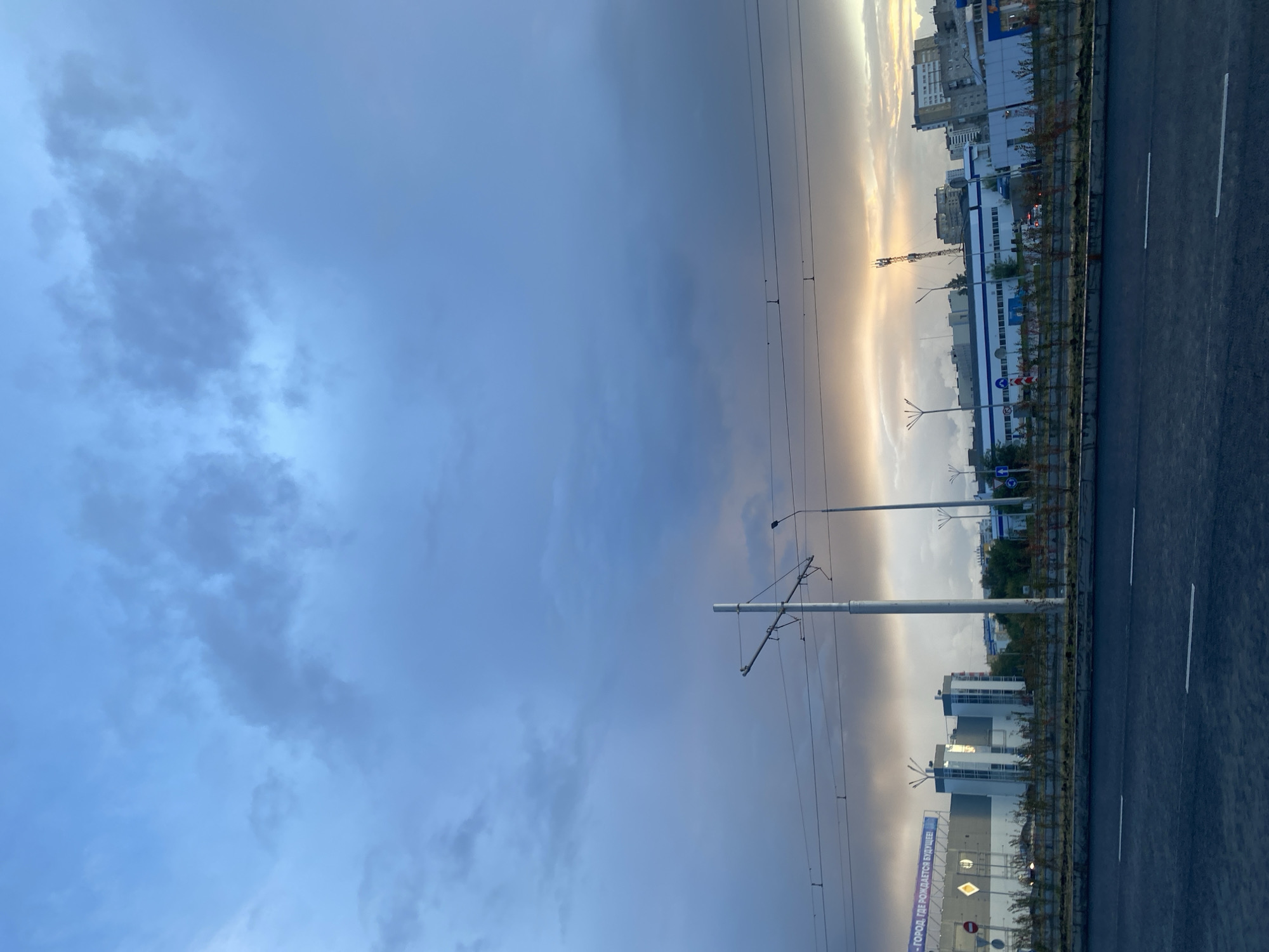 The image captures a street view in Perm, Russia, under a dramatic, cloudy sky. The foreground is dominated by a dark asphalt road with white lane markings. A raised concrete barrier with sparse, small plants separates the road from the sidewalk area. Beyond the barrier, a row of small trees and shrubs lines the edge of what appears to be a construction site or landscaped area.
In the midground, a white pole with overhead tram wires extends upwards, a common sight in urban areas. A street light is visible to the right of the pole. Behind these elements, a modern, multi-story building with a large blue banner bearing Cyrillic text is prominent. The banner reads "ГОРОД, ГДЕ РОЖДАЕТСЯ БУДУЩЕЕ!", which translates to "A CITY WHERE THE FUTURE IS BORN!". Adjacent to this building are other structures, including a white building with blue accents and several taller residential buildings in the background. A telecommunications tower can be seen in the distance.
The sky is filled with a mix of dark, heavy clouds and lighter, illuminated areas, suggesting either an approaching storm or the aftermath of one, with a band of orange-yellow light near the horizon indicating a potential sunset or sunrise. No people or vehicles are clearly identifiable in the frame. The overall impression is of an urban environment at dusk or dawn, with a blend of modern architecture and infrastructure under a dynamic sky.