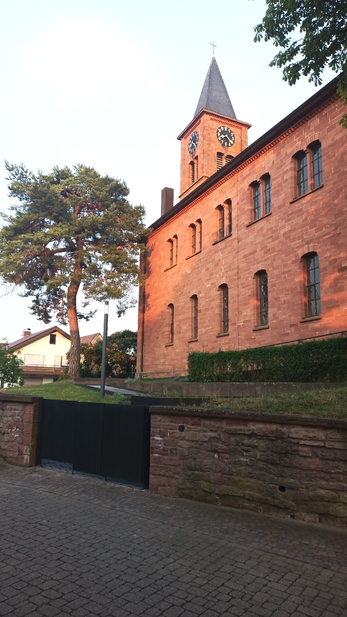 The image shows a church in Epfenbach, Germany, bathed in the warm light of sunset. The church's exterior is constructed from red stone, and its prominent clock tower features two visible clock faces. A tall, mature pine tree stands to the left of the church, casting a shadow. In the foreground, a paved pathway leads towards a black metal gate, which is set into a low stone wall. The sky above is clear and a pale blue, indicating either early morning or late evening.