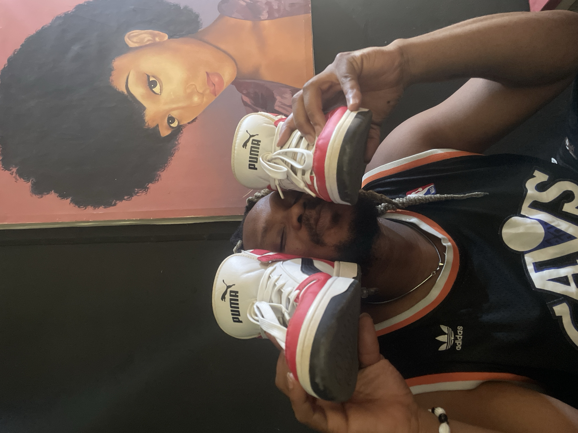 A man wearing a black and orange basketball jersey with an Adidas logo is holding a pair of white, red, and black Puma sneakers up to his face. The background features a painting of a woman with an afro. The location is Jos, Nigeria, as inferred from the prompt.