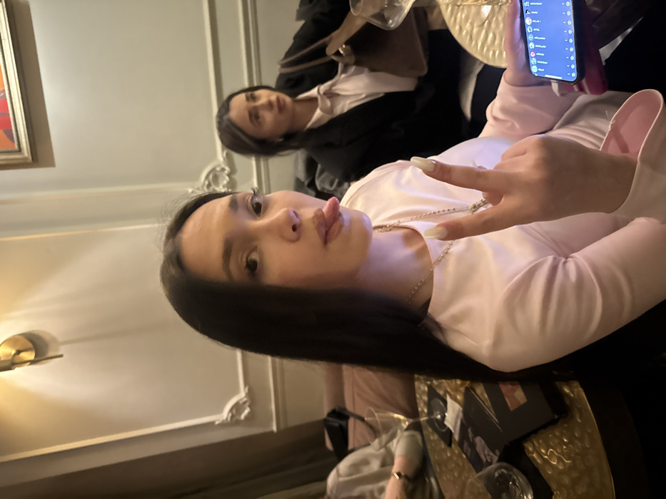 Two women are seated indoors. One is closer to the camera, sticking her tongue out and making a hand gesture while holding a smartphone. She has dark hair and is wearing a light pink top and a beaded necklace. The second woman is further away and appears to be looking at the camera. She has dark hair and is wearing a light pink top with a dark jacket. There are tables with items on them visible in the foreground and background. The interior has ornate molding and wall lighting.