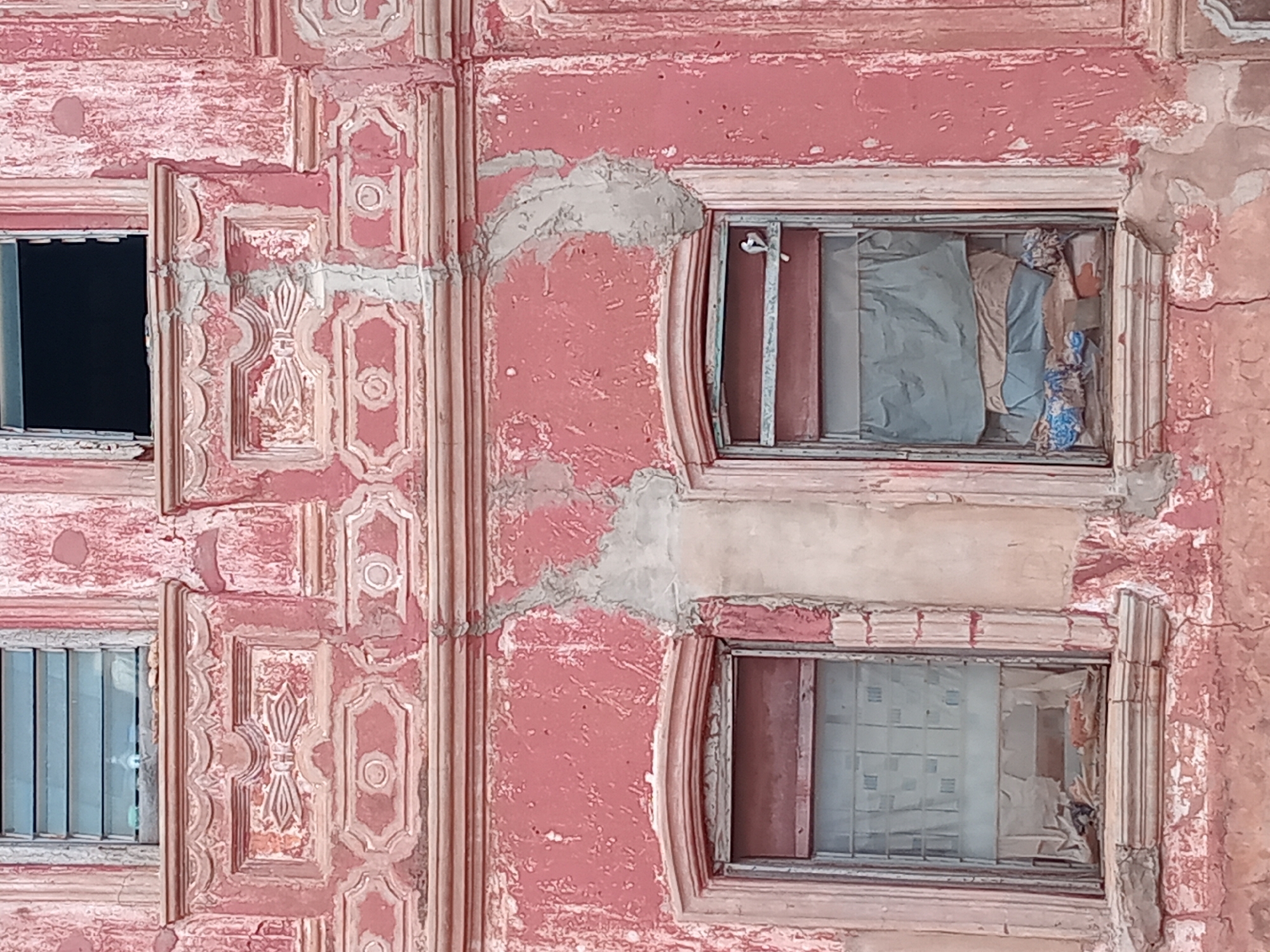 The image depicts a section of a weathered, likely historic, structure in Shagamu, Nigeria. The building features ornate, geometrically patterned architectural details painted in shades of pink and off-white. The paint is heavily chipped and peeling, revealing underlying surfaces and suggesting significant age and lack of recent maintenance. Three windows are visible. The upper left window is dark and appears empty, while the other two windows are partially obscured with interior materials, possibly curtains or other coverings. The structure shows evidence of repair or patching, with areas of newer, gray-colored material filling in damaged sections. The overall impression is one of age, disrepair, and neglect.