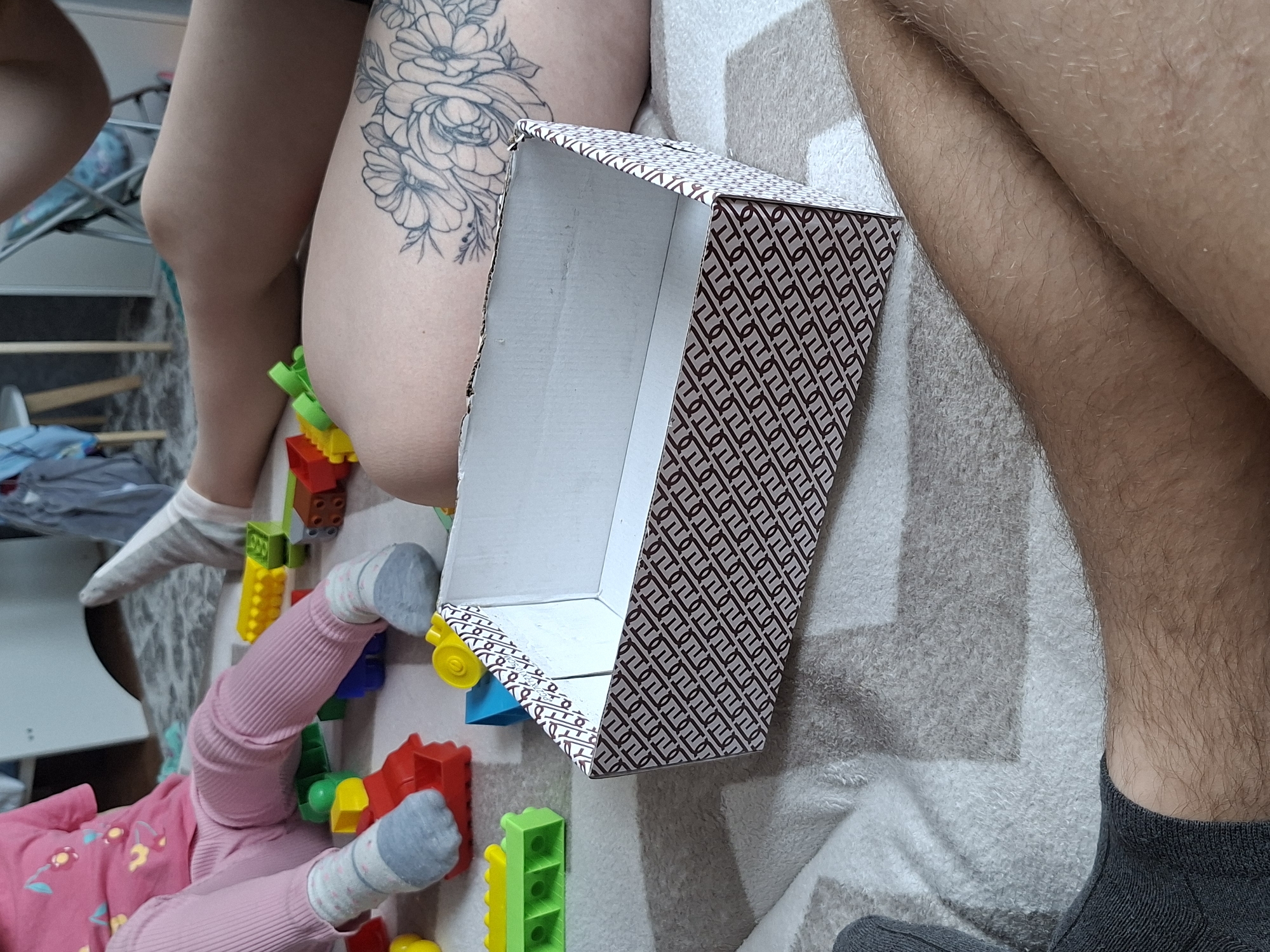 A close-up shot taken indoors depicts an ambiguous scene with parts of people and objects. At the center, there is an open decorative box with a white interior and a patterned exterior. To the left of the box, there is a person's thigh with a floral tattoo, next to it are children's blocks. The bottom portion of the image features a child wearing pink pajamas and gray socks. To the right of the box is the leg of a person in a gray sock. The background is a bed.
