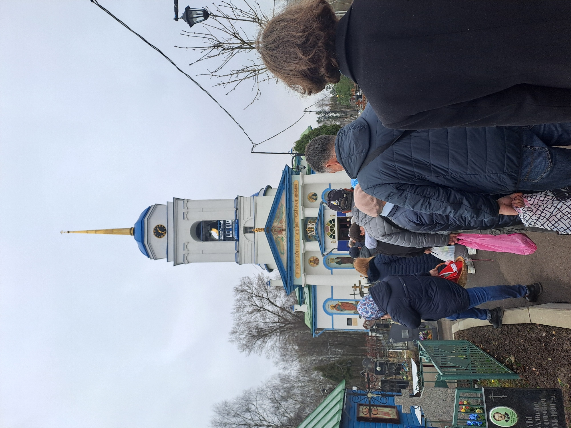 People are gathered outside the Church of the Ascension in Ivanisovo, Russia. The church is white with blue trim and a gold spire. The people are dressed in warm clothing. Some are holding bags and baskets. The scene is likely taking place in a cemetery as there are gravestones and memorials visible around the church. The sky is overcast.