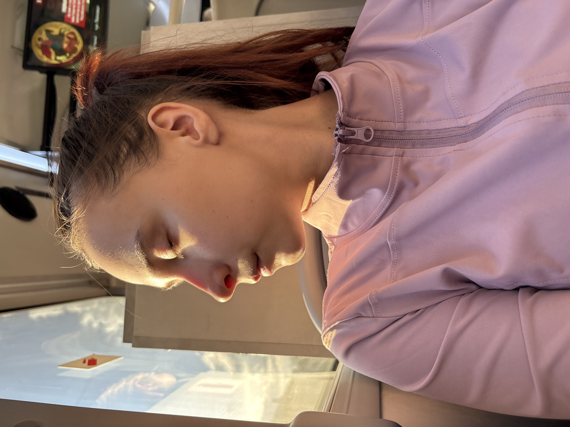 A woman with dark hair in a high ponytail and a pink jacket is looking down at a surface, potentially a display or table. The setting appears to be indoors, with a display or screen partially visible in the background. The lighting is bright, creating highlights on the woman's face. A small object on a light table is also visible. The location in the city of Perm, Russia, is not discernible from the image.