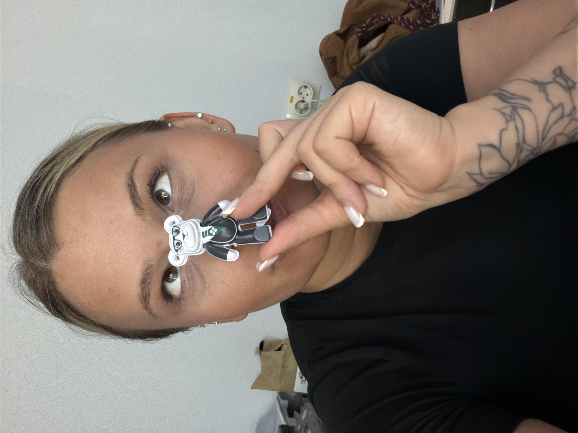 A fair-skinned person with brown eyes, light brown hair with blonde highlights, and multiple piercings in their left ear is holding a small, flat, cartoon bear figurine (black and white with a green accent on its chest) in front of their face, near their mouth. The person is wearing a black shirt and has a tattoo of a stylized floral design on their right forearm. The background is a light gray wall with an electrical outlet and a brown bag visible. The image is rotated counter-clockwise, making the orientation appear sideways. The location is not definitively identifiable as Sarapul, Russia based on the provided image.