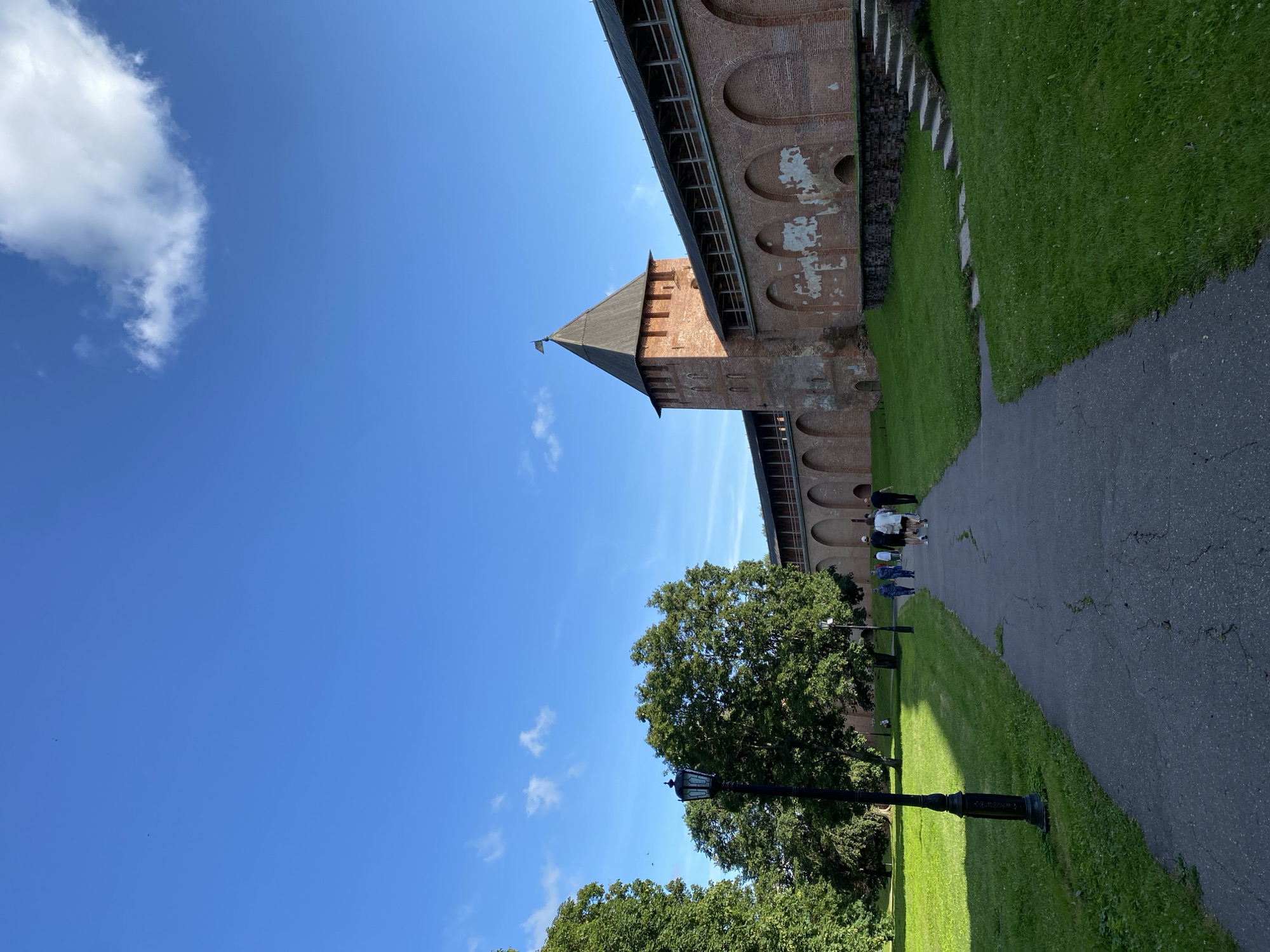 A paved pathway leads through a grassy park towards a large, ancient brick fortification with a prominent tower. Several people are walking away from the camera on the pathway, and some are further up the path closer to the fortification. The fortification is made of red brick, with arched openings visible along its length and a wooden walkway on top. The tower is also brick, with a dark, pointed roof and a small flag at its apex. Tall, green trees are on the left side of the pathway, and a black lamppost stands near the foreground. The sky is bright blue with a few wispy clouds. This scene is located at the Novgorod Kremlin in Veliky Novgorod, Russia, a historic fortress complex.