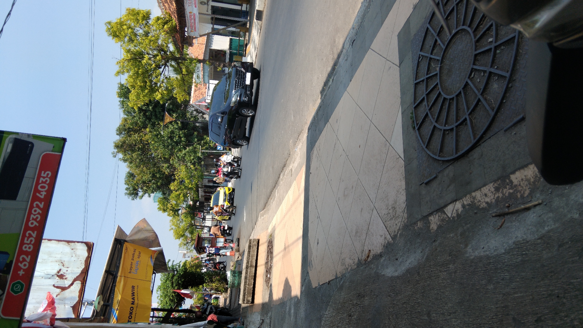 This image depicts a street scene in Batang, Indonesia, likely near a commercial area. In the foreground, a sidewalk with square tiles transitions to a darker, textured surface with a circular metal grate. A black motorcycle is partially visible in the bottom right corner. The street itself is paved and shows signs of traffic. Several vehicles are present, including a black SUV, a yellow car, and multiple motorcycles, some with riders. People are seen walking on the sidewalk and riding motorcycles. Buildings line the right side of the street, some with awnings and visible signage. A large green tree with yellowing leaves stands prominently in the middle ground, with other trees and vegetation visible. Overhead, a blue sky with faint clouds suggests a sunny day. A red and green sign with white text, including a phone number "+62 852 9392 4035", is mounted on a pole on the left side of the image. Another yellow banner with Indonesian text and a logo is also visible on the left. The overall impression is a bustling urban environment.