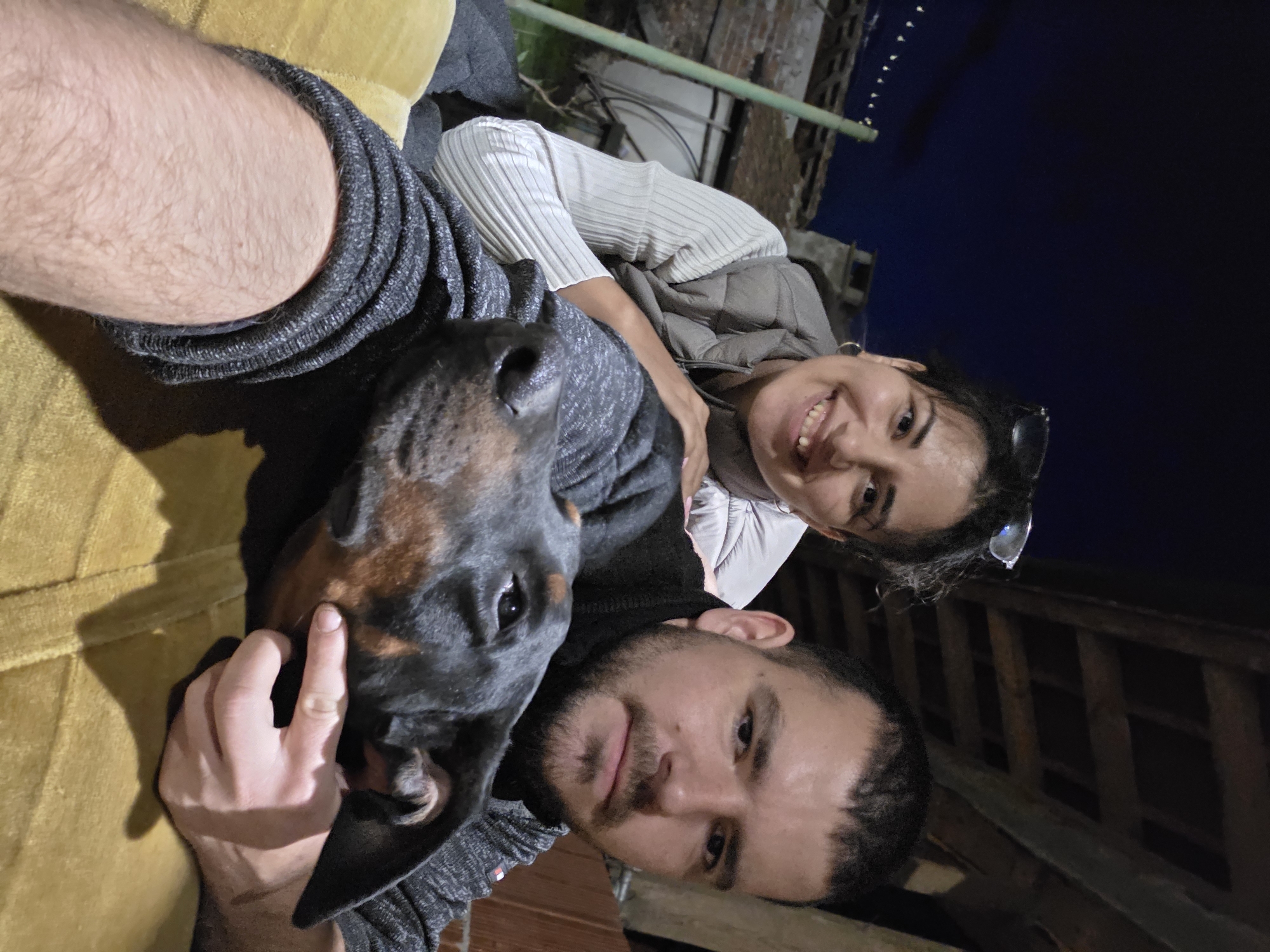 A selfie-style photograph taken at night depicts two adults and a Doberman Pinscher. A female subject, identifiable as an adult with dark hair, glasses on her head, a light-colored ribbed long-sleeved top, and a grey puffer vest, smiles directly at the camera from the left, positioned behind a male subject. Her right hand rests on his shoulder. The male subject, an adult with short dark hair and a beard, occupies the right foreground, holding the camera. He wears a dark long-sleeved shirt/sweater, possibly with a black turtleneck. His left hand gently touches the muzzle of a black and tan Doberman Pinscher, whose head is prominent in the lower center, resting on a textured yellow surface. The background shows a dark night sky in the upper left, with distant string lights and a vertical green metal pole. Behind the subjects, a partial view of light-colored brick structures, a chimney-like element, and dark wooden beams or rafters are visible, suggesting an outdoor or semi-outdoor setting such as a patio or backyard. The specific location within the city of Unknown, Unknown is not discernible.