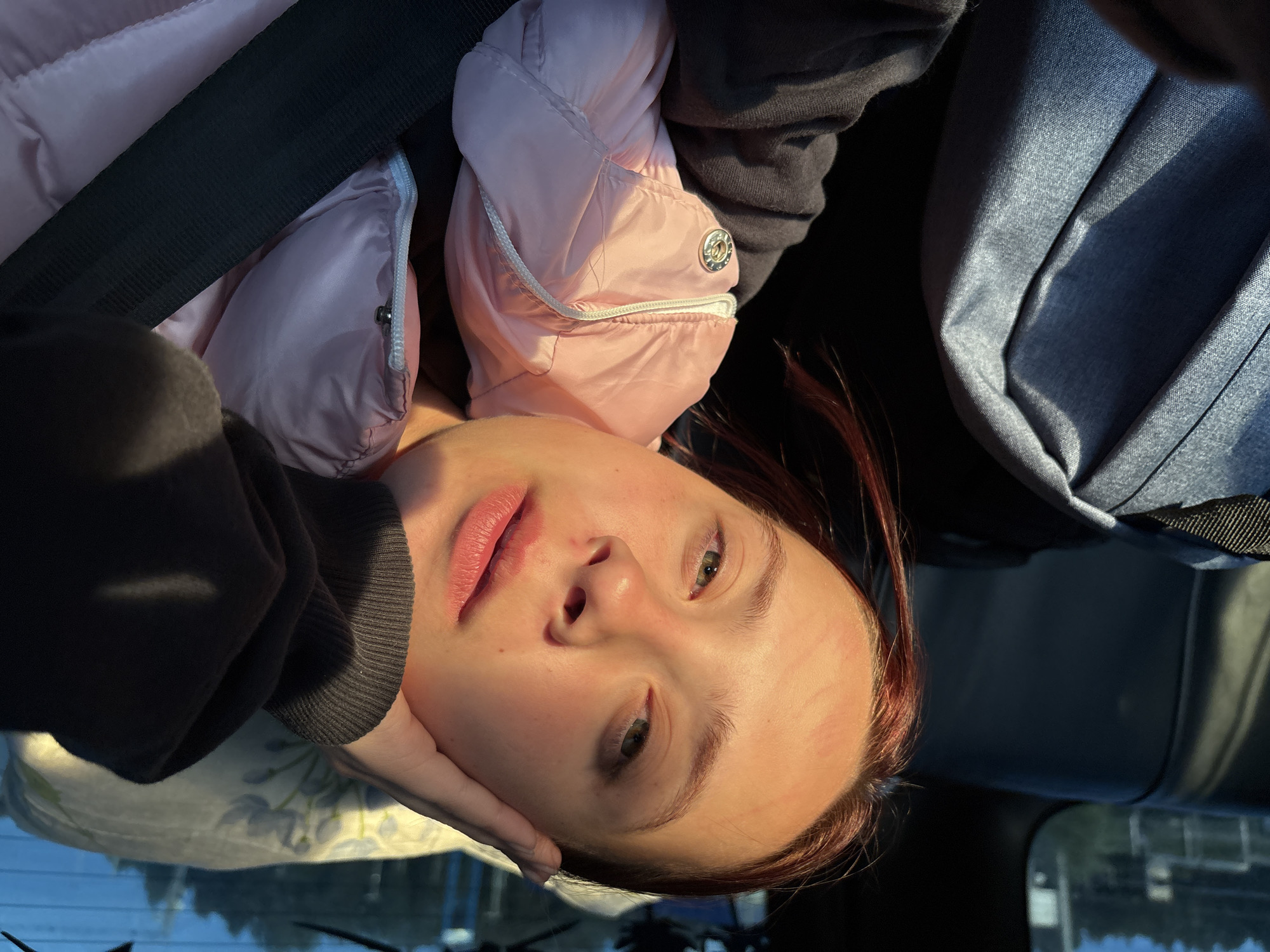 A person with light skin and red hair is in the passenger seat of a vehicle. She is wearing a pink puffy jacket and a black hooded sweatshirt. She is wearing a seatbelt. The person's left hand is touching her cheek. Behind her to the right, out of focus, is a blue-grey bag. The windshield shows the sky and a blur of trees and buildings outside. Based on the prompt stating location, it is Прикамский, Russia.