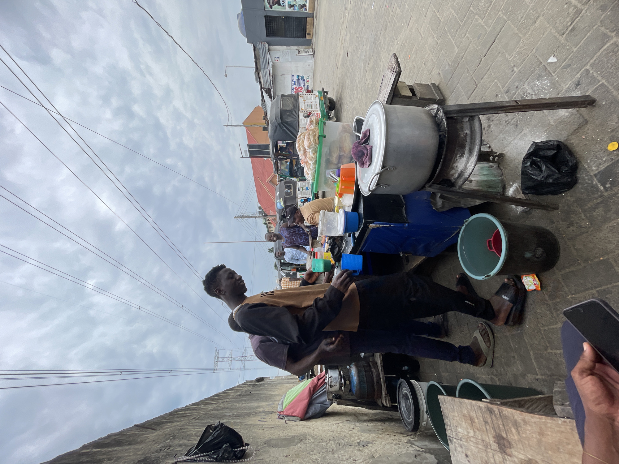 The image shows a street food vendor in Ado, Nigeria. A dark-skinned man in a brown and black long-sleeved shirt and black pants is standing near a cooking setup consisting of a large metal pot on a makeshift stove constructed from metal rims and wood. The cooking station includes various containers, buckets, and utensils. Customers are visible in the background. Overhead power lines crisscross the sky. The ground is paved, and buildings are visible in the background, indicating an urban setting.