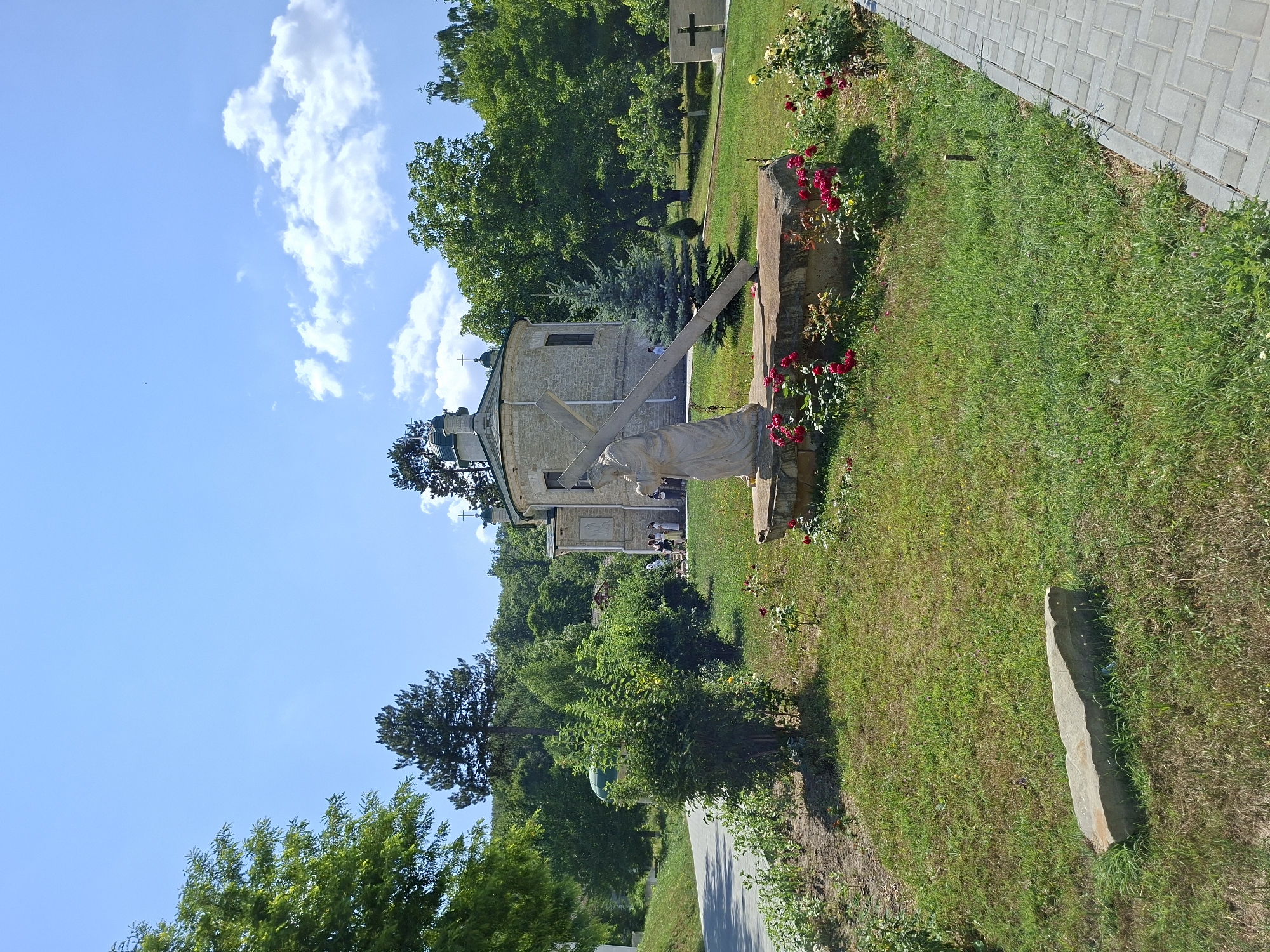 The image depicts an outdoor scene with a religious theme at the Condrița Monastery in Moldova. In the foreground, a life-size stone sculpture of Jesus carrying the cross is positioned on a stone base, surrounded by red roses. To the right, a paved walkway runs along a grassy area. In the mid-ground, a light-colored, rounded church with a green dome is visible. Several individuals, some wearing religious attire, are standing near the church entrance. A large tree stands behind the church, and a dense line of trees forms the background. The sky is blue with scattered white clouds.