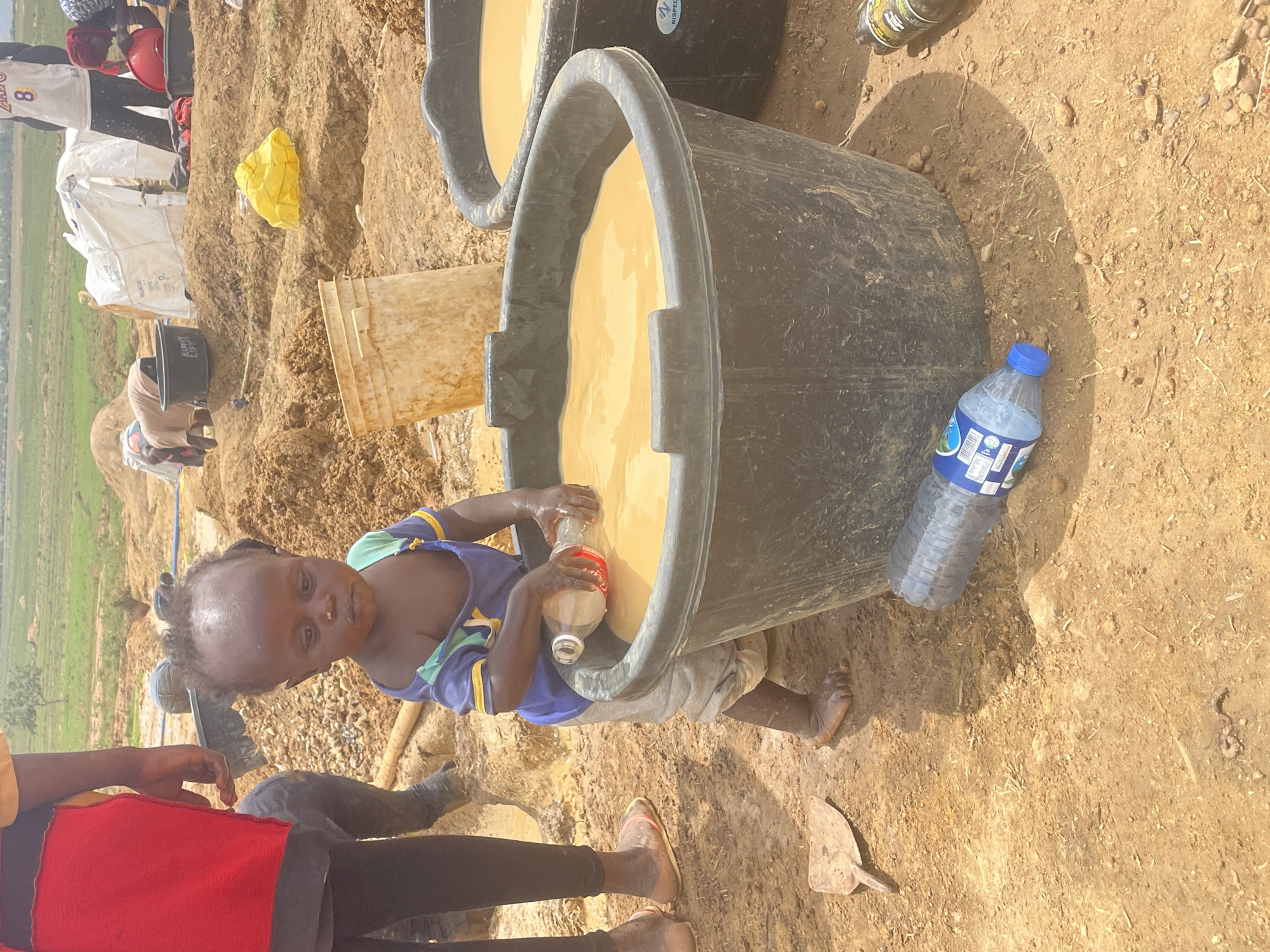 A photograph depicts a child filling a bottle with liquid from a large black tub in what appears to be an outdoor setting, likely in Durumi, Nigeria. The child, wearing a blue and green top and light-colored pants, is standing beside the tub, which is filled with a beige-colored liquid. A second, similar tub is partially visible behind the first. A bottle of water rests on the ground next to the tub. Other individuals are partially visible in the image, with legs and feet visible in the foreground. The ground is dry and dusty. In the background, there's a landscape with green fields.