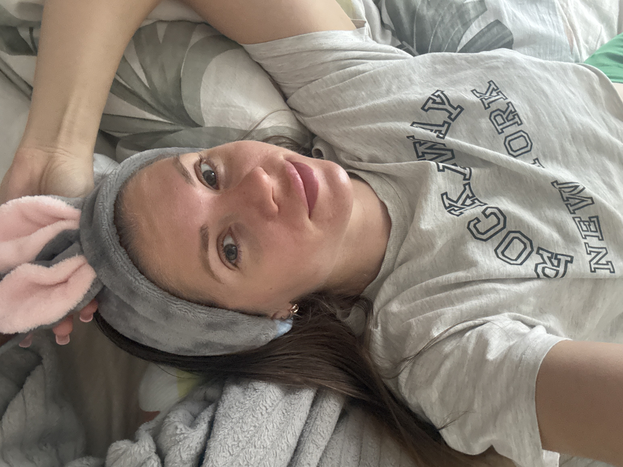 A woman lies in bed wearing a gray headband with pink bunny ears. She is wearing a gray t-shirt with "ROCKY NEW YORK" printed on it. She is lying on a gray and white patterned bedspread with a gray fleece blanket partially covering her. The location is indiscernible without further information.