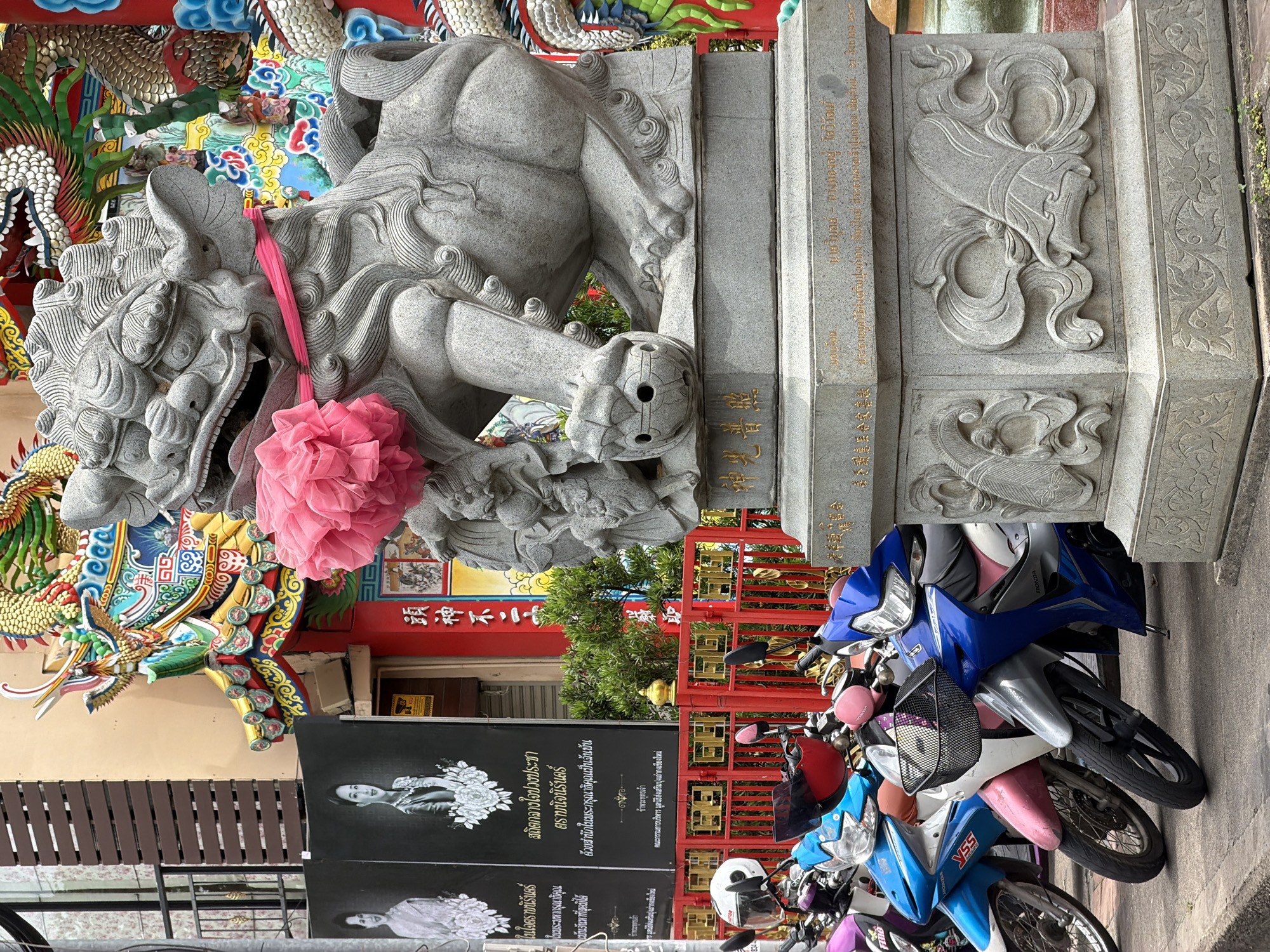 This image, taken in Chiang Mai City Municipality, Thailand, presents a scene with a blend of religious and cultural elements, alongside everyday life. Dominating the foreground is a large, ornate stone carving, likely a guardian lion or similar mythical creature, adorned with a bright pink fabric flower and ribbon. Behind it, a vibrant, colorful mural depicts a dragon, typical of Chinese or Thai-Chinese temples and shrines.
To the right, a collection of motorcycles are parked, indicating a public space or a place of gathering. The motorcycles, in various colors including blue, pink, and white, suggest a modern, urban environment. In the background, a red gate with golden embellishments and greenery peeking through add to the visual richness.
A black banner with images of a woman and floral motifs is displayed prominently. The banner contains Thai text, with some legible phrases like "ในใจตราบนิรันดร์" (In the heart forever) and "สถิตกลางใจปวงประชา" (Resides in the heart of the people), suggesting a memorial or tribute. Other text on the banner, in Chinese characters, includes "頭神不二" (Head God Not Two), further reinforcing the cultural context.
The overall impression is one of a cultural landmark or a temple area, possibly during a festival or a day of remembrance, where traditional artistry coexists with the daily routines of the local community. The lighting suggests it might be daytime, with diffused sunlight.