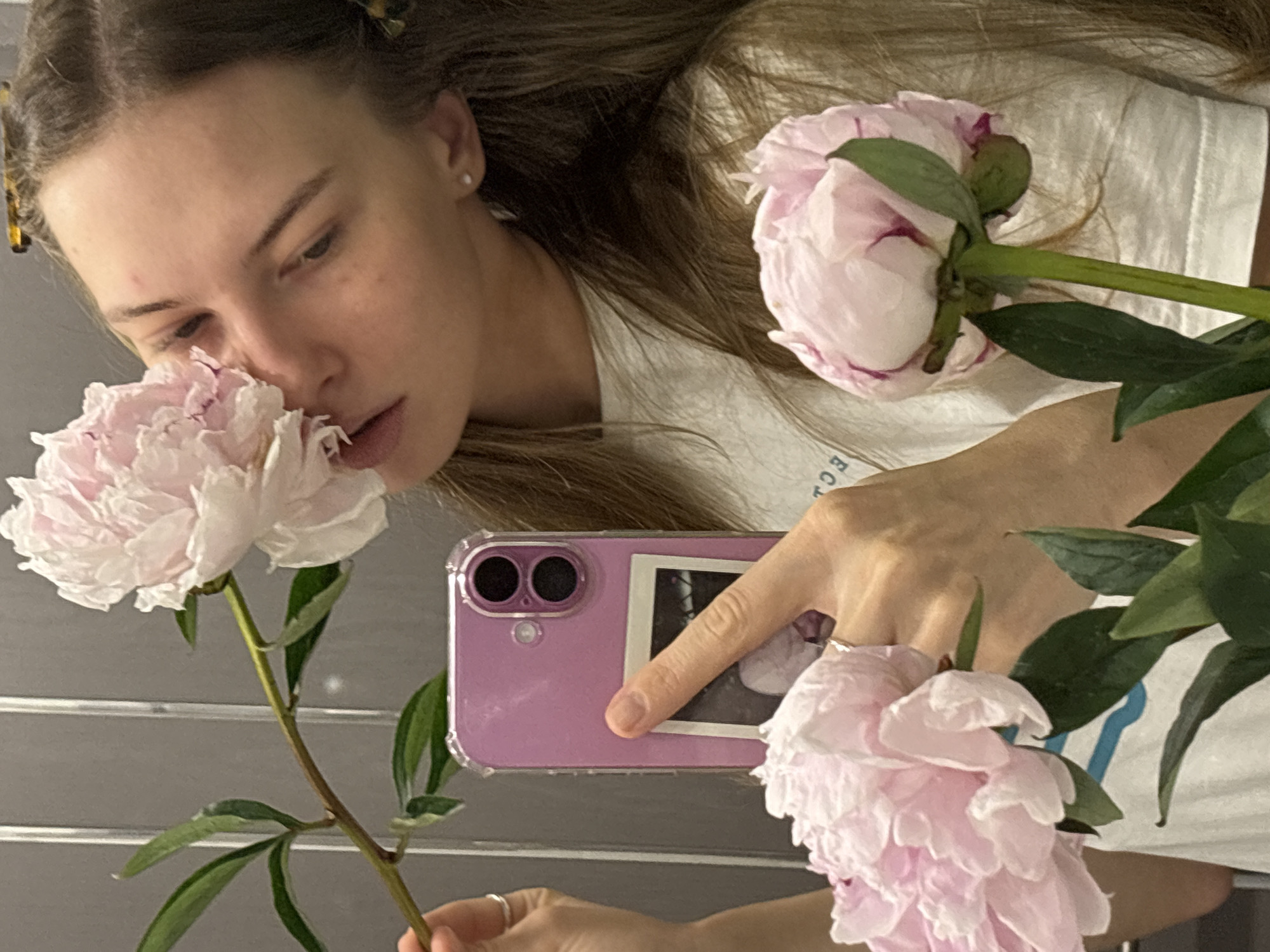 The image is a selfie taken in what appears to be a bathroom with grey tiled walls. A young woman with long brown hair and a white t-shirt is holding a bouquet of pink peonies. She is bringing one of the flowers close to her face, almost touching her nose with it. In her other hand, she is holding a pink phone with a clear case, in which a small polaroid photo is stuck. A second peony is visible at the bottom right of the frame. The woman has a light complexion with freckles and is wearing a small stud earring. The lighting is soft, creating a natural and intimate atmosphere. The scene does not provide any specific clues to identify the location as Perm, Russia.
