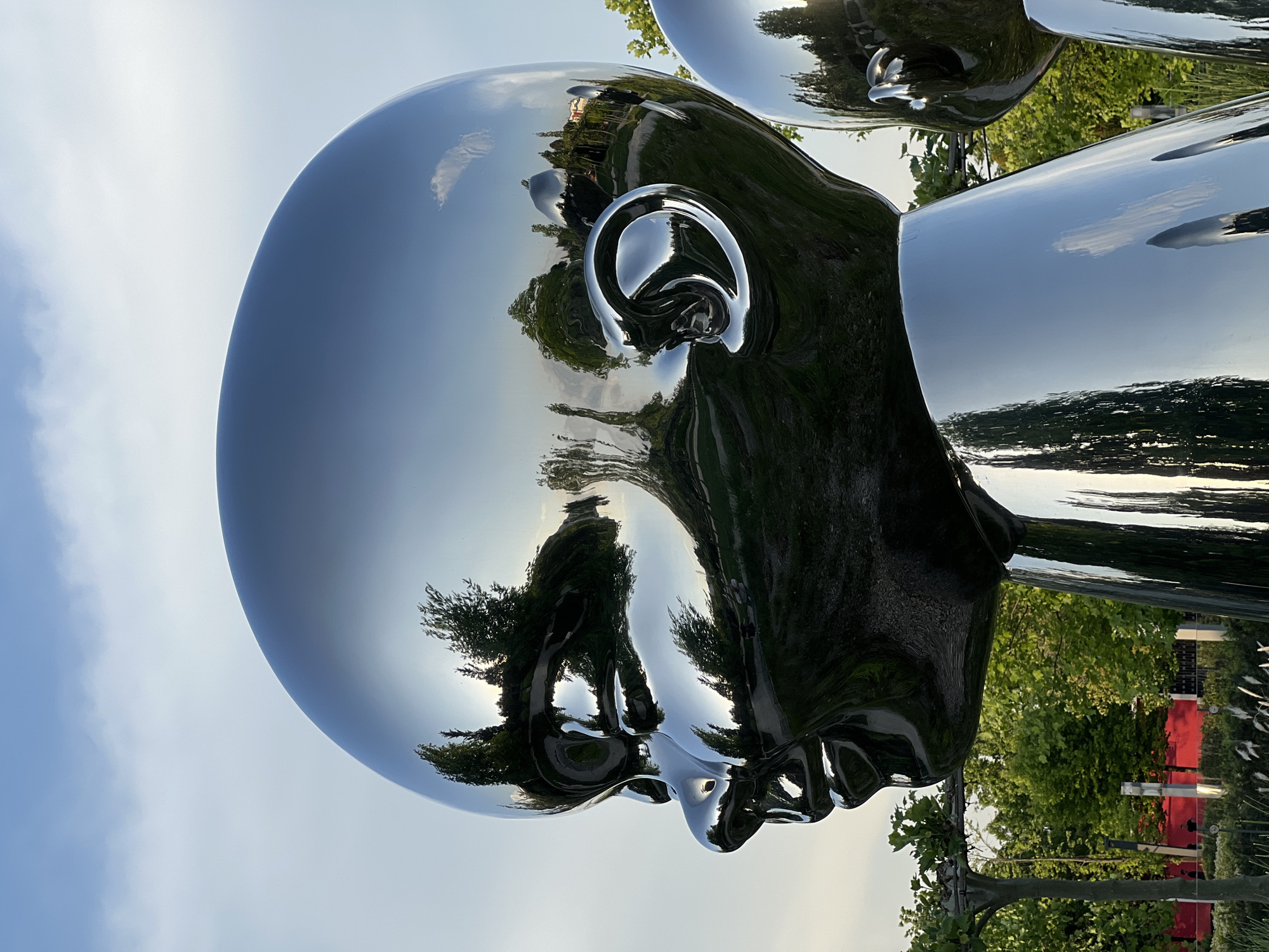 The image depicts a shiny, metallic sculpture of a human head in Krasnodar, Russia. The sculpture's surface reflects the surrounding environment, including trees, sky, and other parts of the sculpture itself. The head is positioned in profile, with the ear clearly visible. The reflection creates a distorted and abstract view of the surroundings. Another metallic head is visible in the background, partially out of focus.
