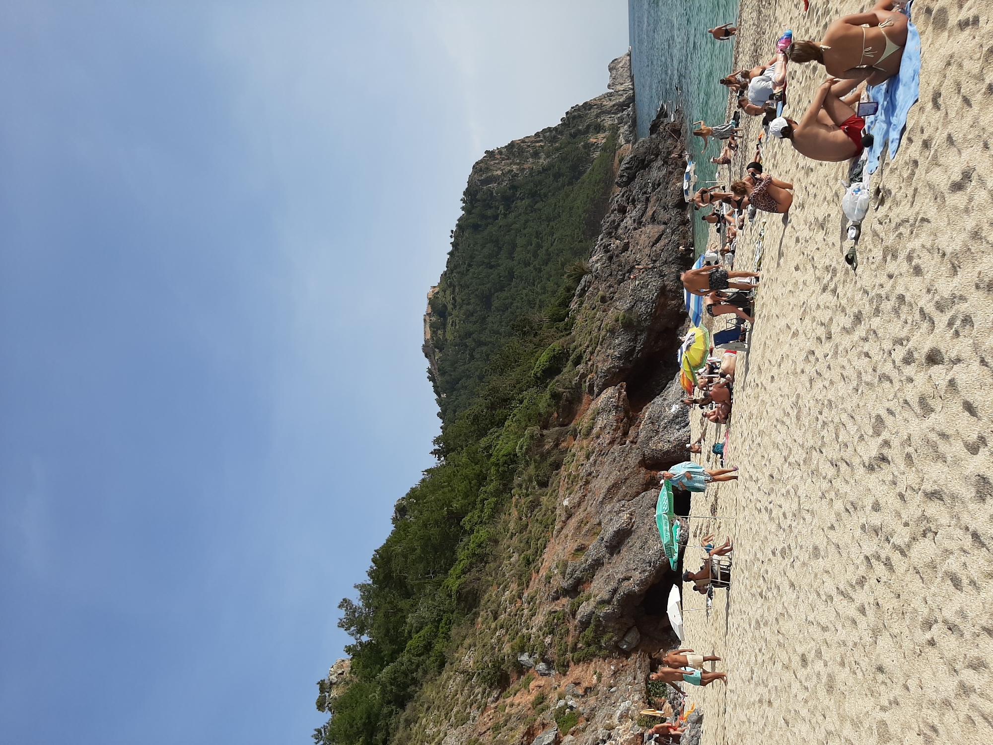 The image captures a sunny beach scene in Alanya, Turkey. A large rock formation covered in greenery dominates the left side of the frame, transitioning into rocky cliffs along the water's edge. The beach is populated with numerous people sunbathing, swimming, and relaxing under umbrellas. Individuals can be seen in swimsuits and casual attire. The blue sea stretches out from the beach, meeting a clear blue sky. The overall atmosphere suggests a warm, sunny day at a popular tourist destination.