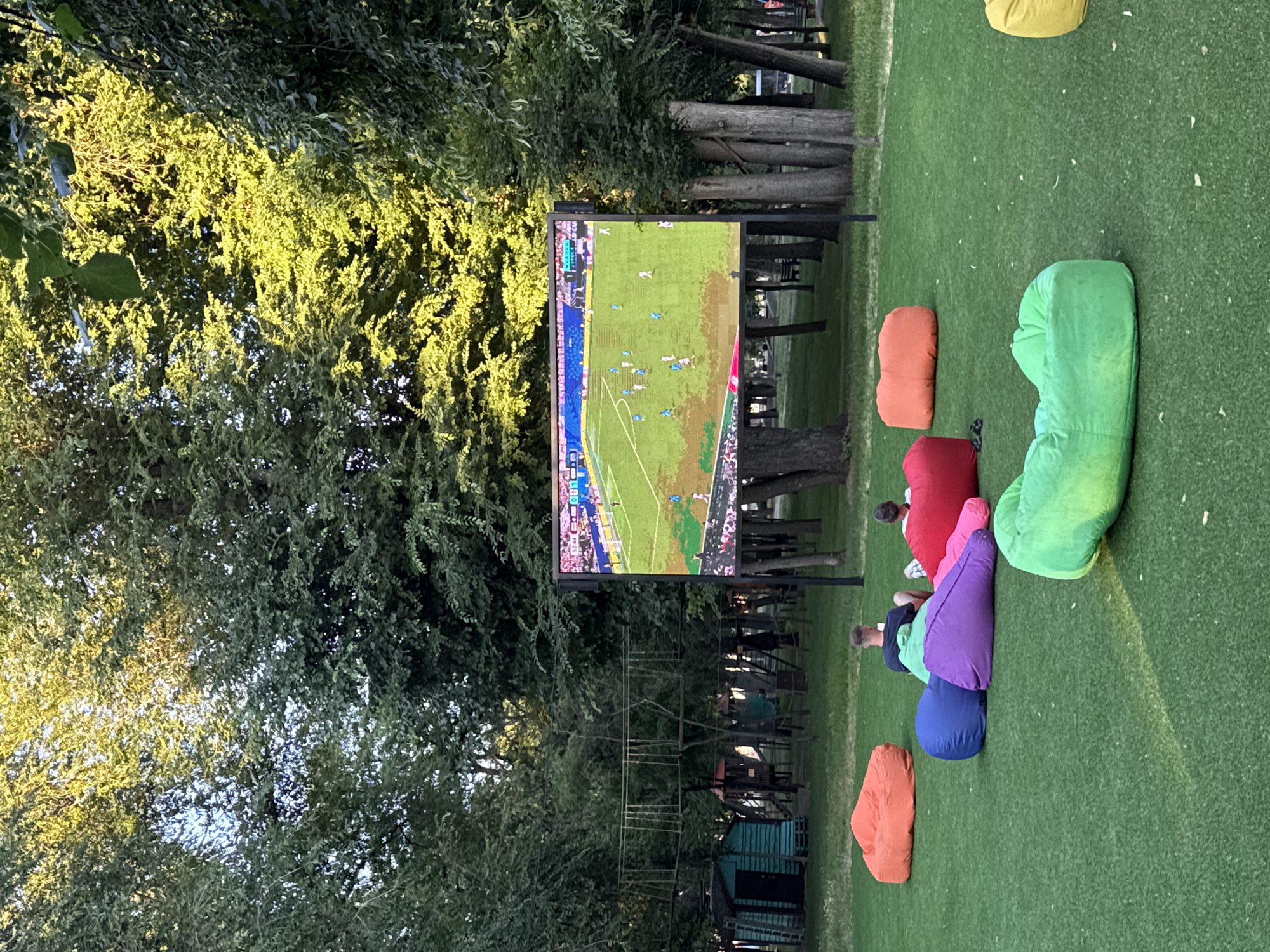 The image depicts an outdoor screen displaying a soccer match in a park-like setting. A group of people are seated on colorful beanbag chairs arranged on artificial turf. In the background, trees and playground equipment are visible. The scene suggests a casual gathering for watching sports in an urban green space. The specific location in Dnipro, Ukraine, cannot be determined from the image alone.