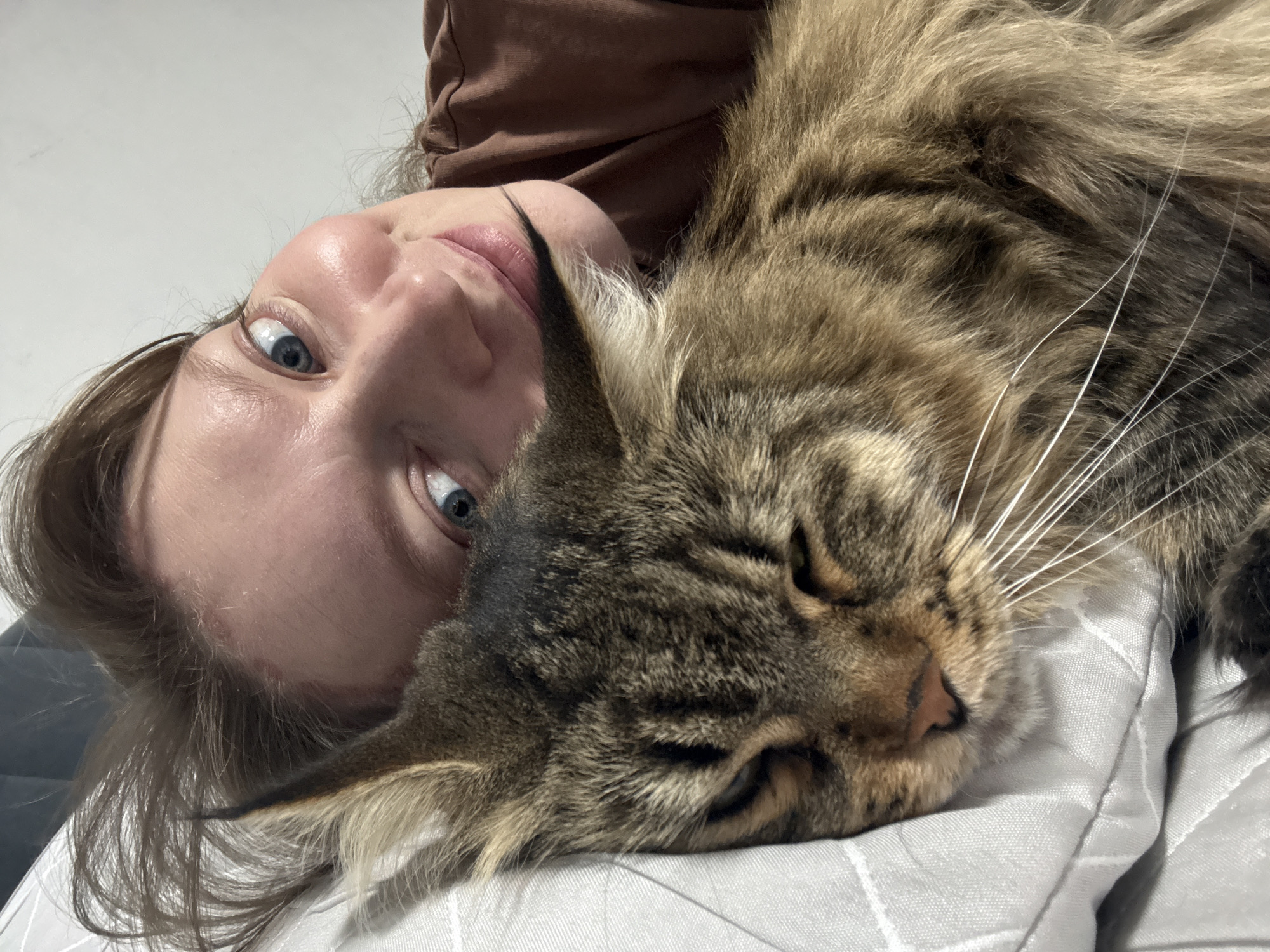 A woman with fair skin and blue eyes is lying on her back, looking at the camera. Her head is tilted to the left. Her brown hair is parted to the side. She is wearing a brown shirt. A large, brown tabby cat is lying on top of her. The cat's head is resting on a white pillow with a stitched square pattern. The location in the city of Izhevsk, Russia is unknown.