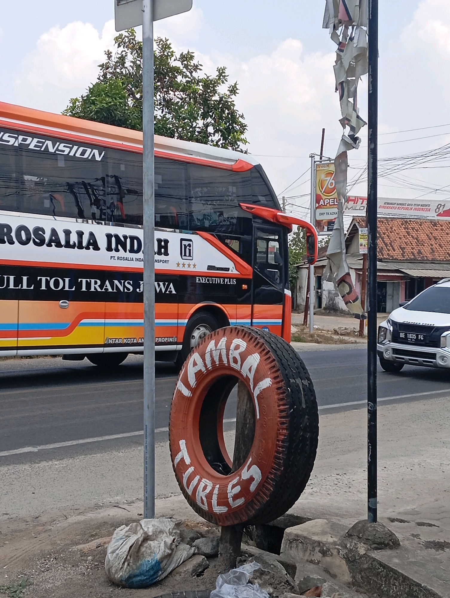 The image depicts a street scene in Ganjar Asri, Indonesia. A large orange and white bus, identified as "ROSALIA INDAH TRANSPORT," is parked on the left side of the frame, with the text "FULL TOL TRANS JAWA" visible on its side. In the foreground, a painted tire, possibly repurposed as a roadside decoration, is prominently displayed, with white lettering that appears to spell out "LAMBAL TURLES." A white SUV with license plate "BE 1835 FG" is visible on the right, driving on the road. Various signs and buildings are visible in the background, suggesting an urban or semi-urban setting. A crumpled plastic bag lies on the ground near the tire. The sky is overcast with clouds.