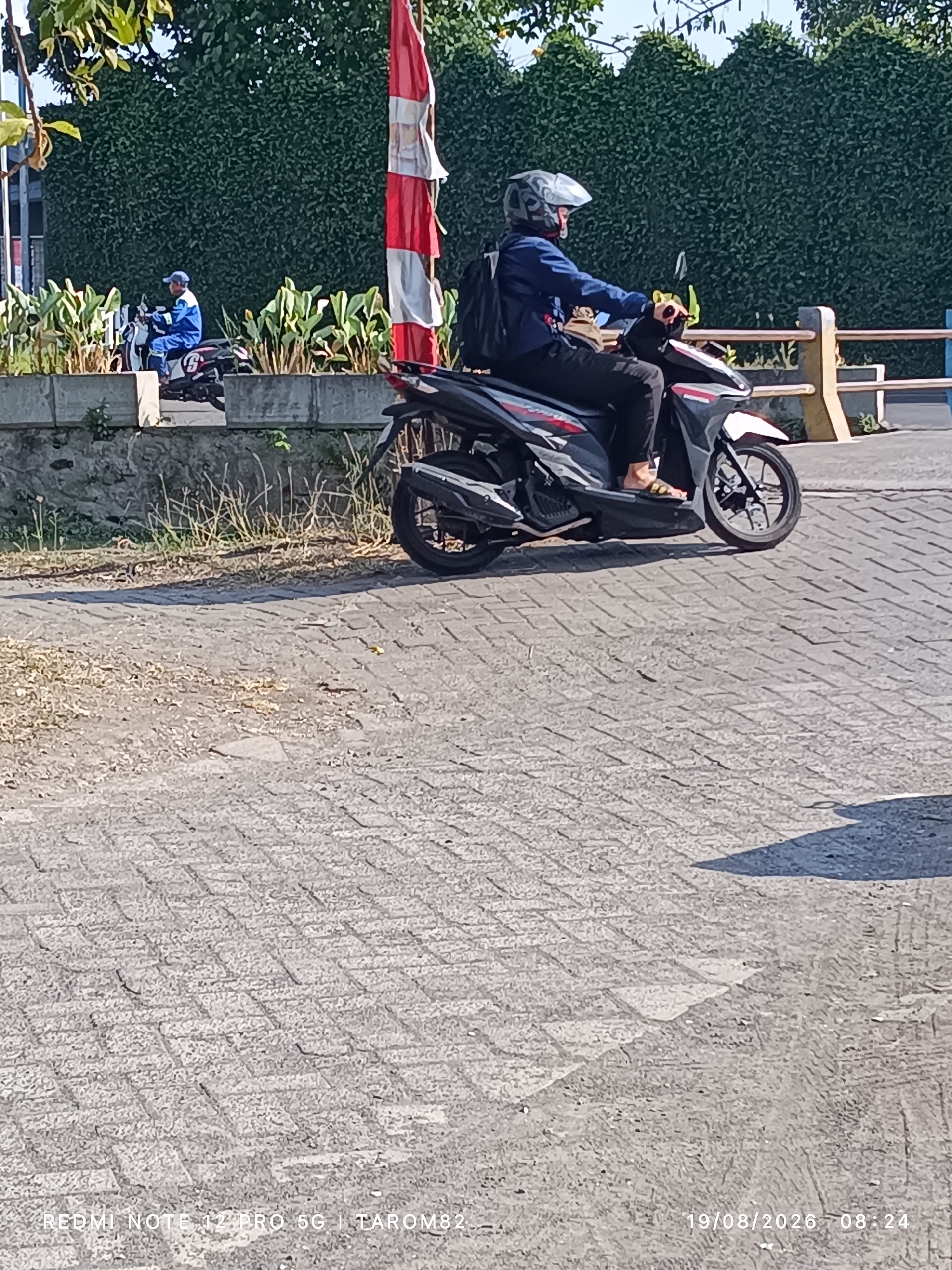 In the city of Tebel, Indonesia, a person is riding a scooter on a paved street. The rider, wearing a blue jacket and a helmet, is positioned in the center of the frame. The scooter is a dark color with red and white accents. In the background, there is a dense green hedge and a red and white flag. The ground is paved with interlocking bricks. The timestamp in the lower right corner indicates the photo was taken on August 19, 2026, at 8:24 AM.